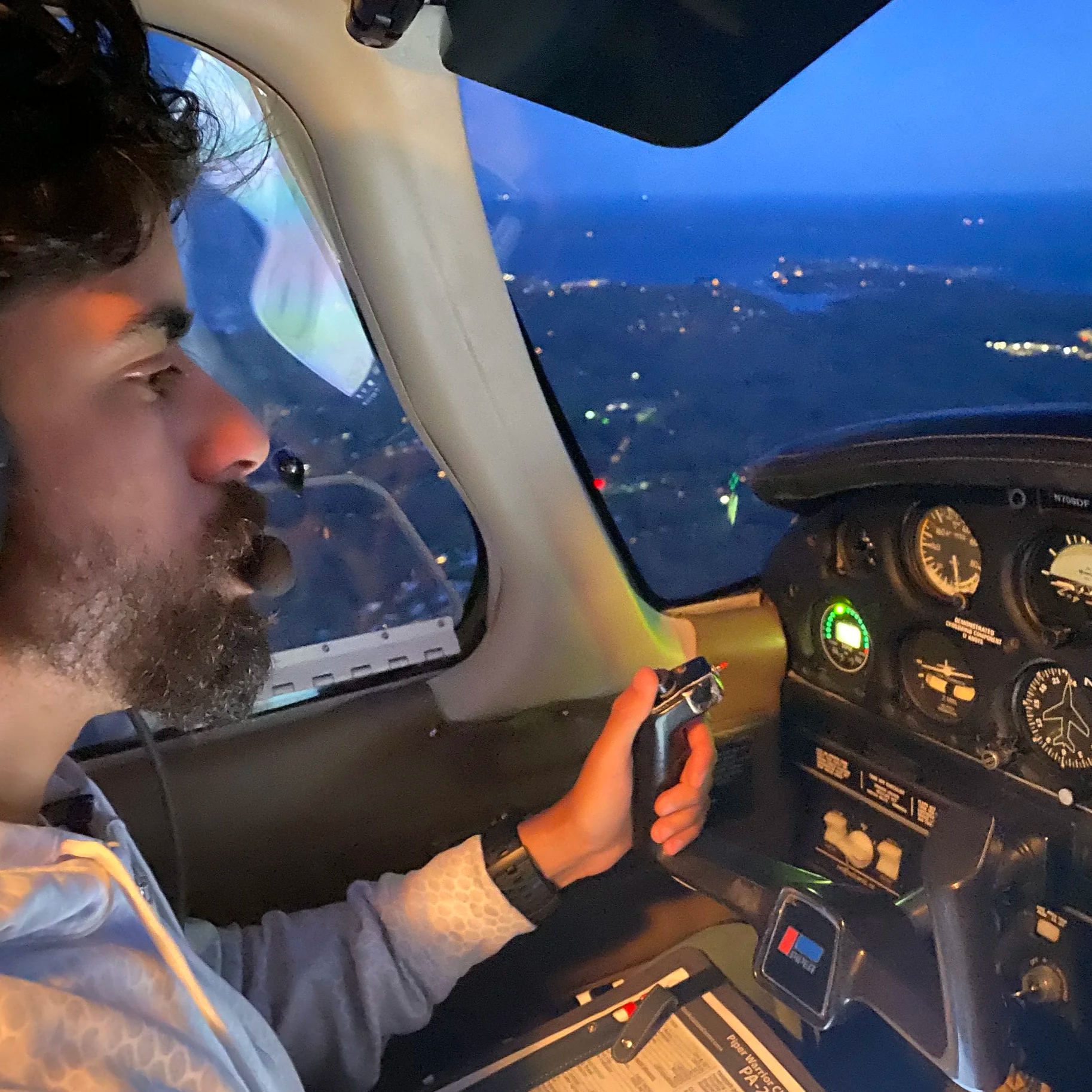 A man with dark hair and a beard sitting in the cockpit of a small aircraft during dusk, holding a control device, with various flight instruments and a view of the landscape below through the window.