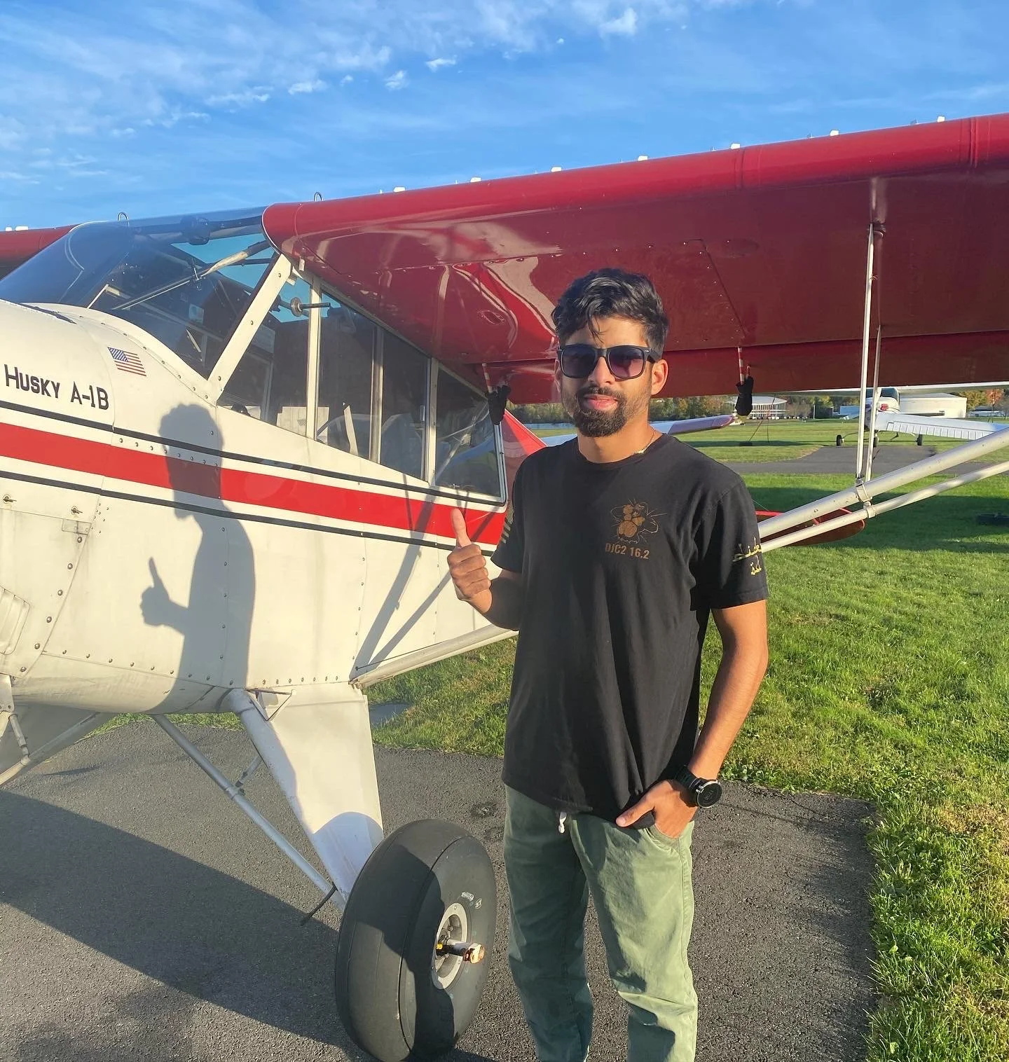 A man in sunglasses giving a thumbs-up standing next to a small white and red airplane on a grassy field on a sunny day.