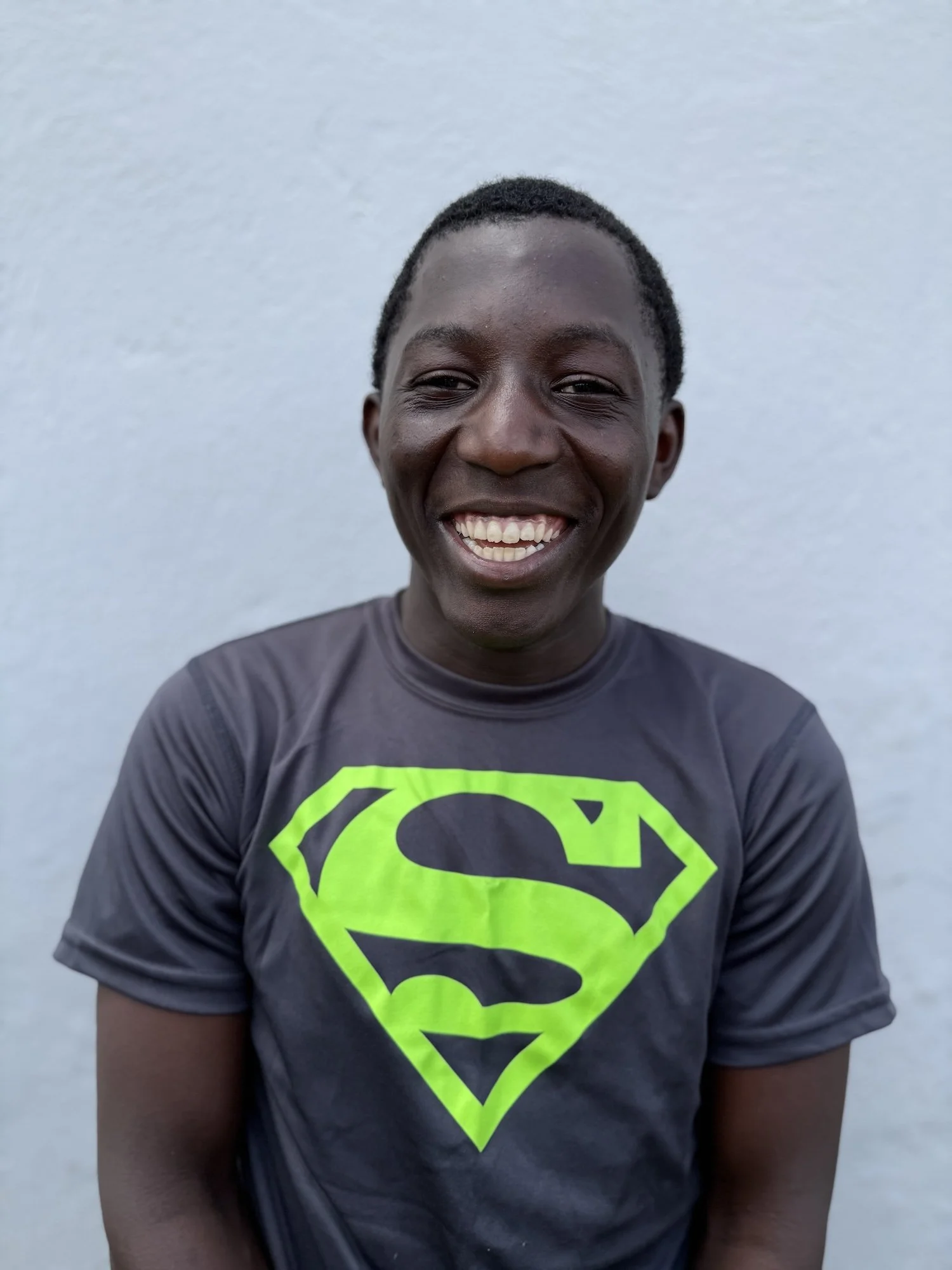 A smiling young person with dark skin and short curly hair wearing a black T-shirt with a bright green Superman logo, standing in front of a plain light-colored wall.