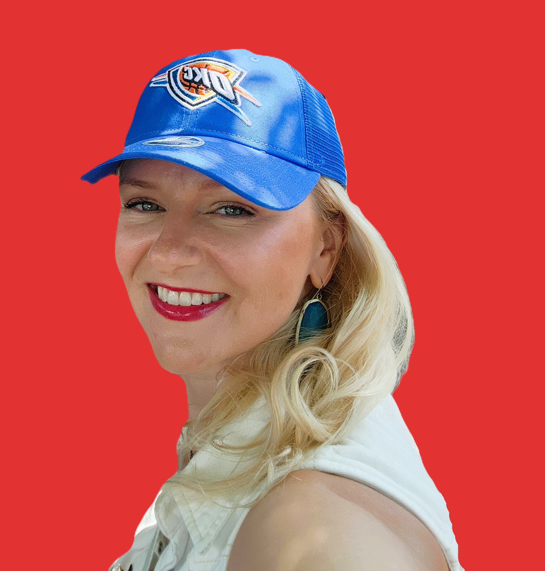Portrait of a woman with blonde hair, wearing a blue baseball cap with a logo, earrings, red lipstick, and a light-colored sleeveless top, smiling against a red background.