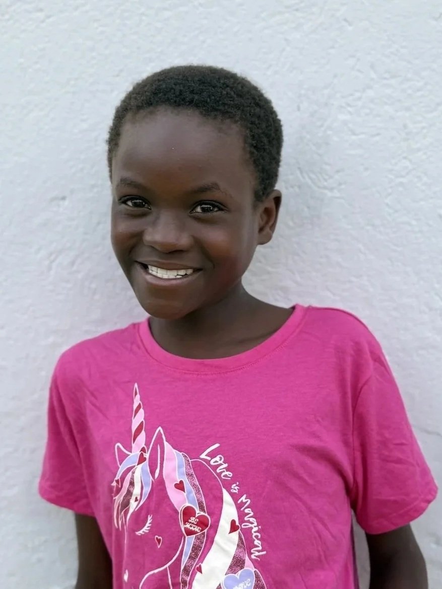 A smiling girl with short curly hair, wearing a pink t-shirt with a unicorn graphic and the words 'Love is Magical,' standing against a textured white wall.