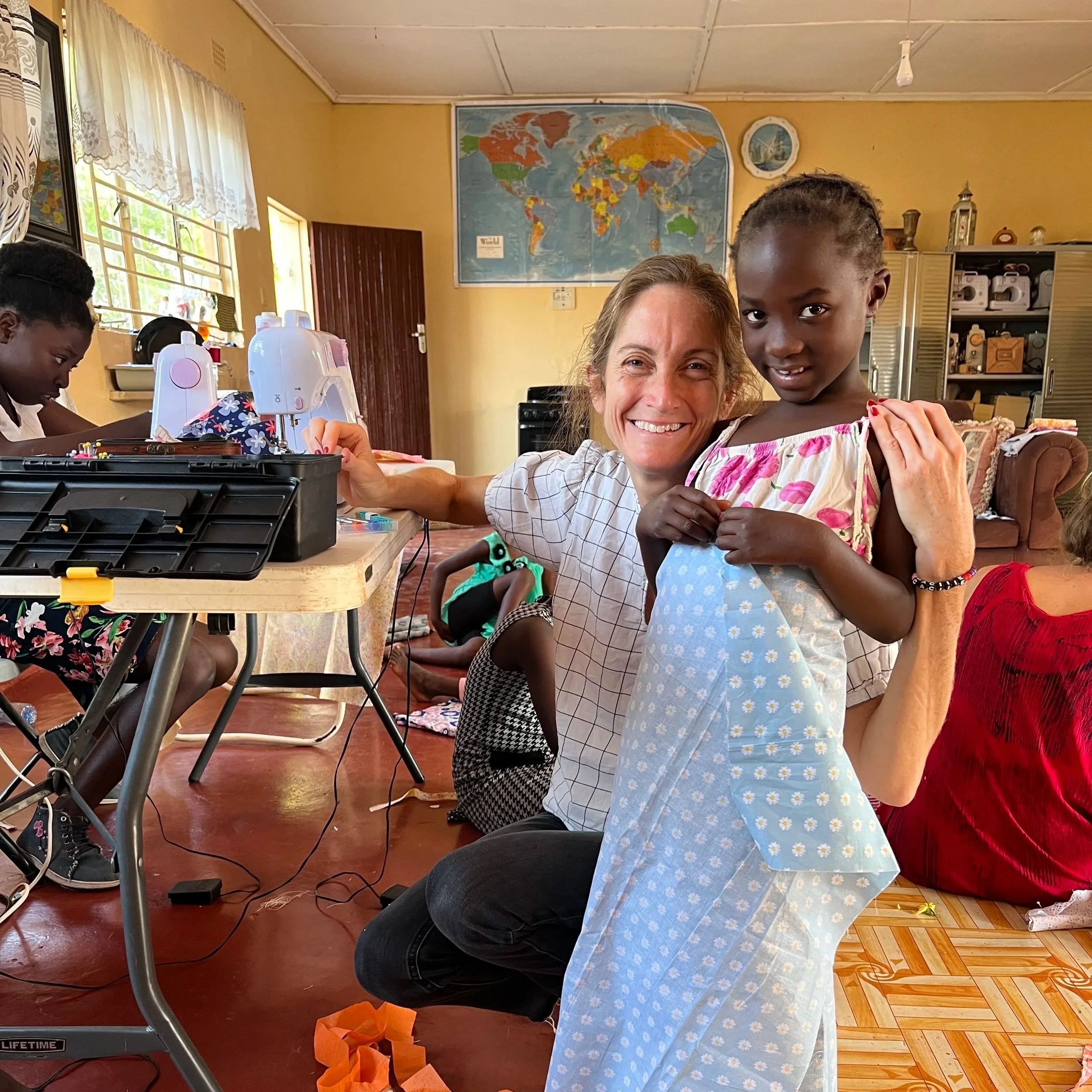A smiling woman and young girl posing together indoors, with sewing machines and other children in the background.
