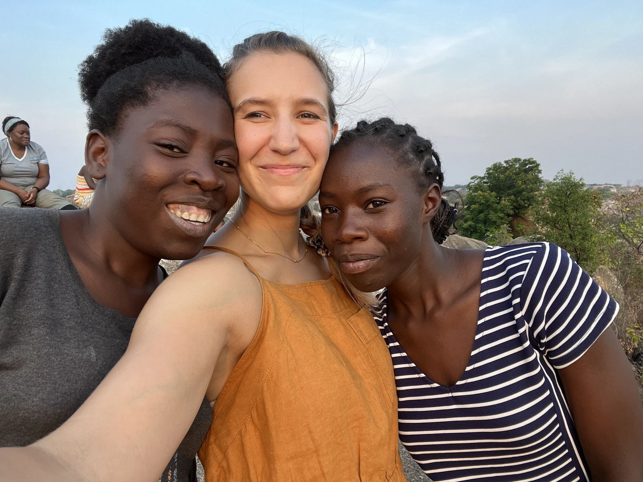 Three women taking a selfie outdoors, smiling, with a woman sitting in the background and trees in the distance.