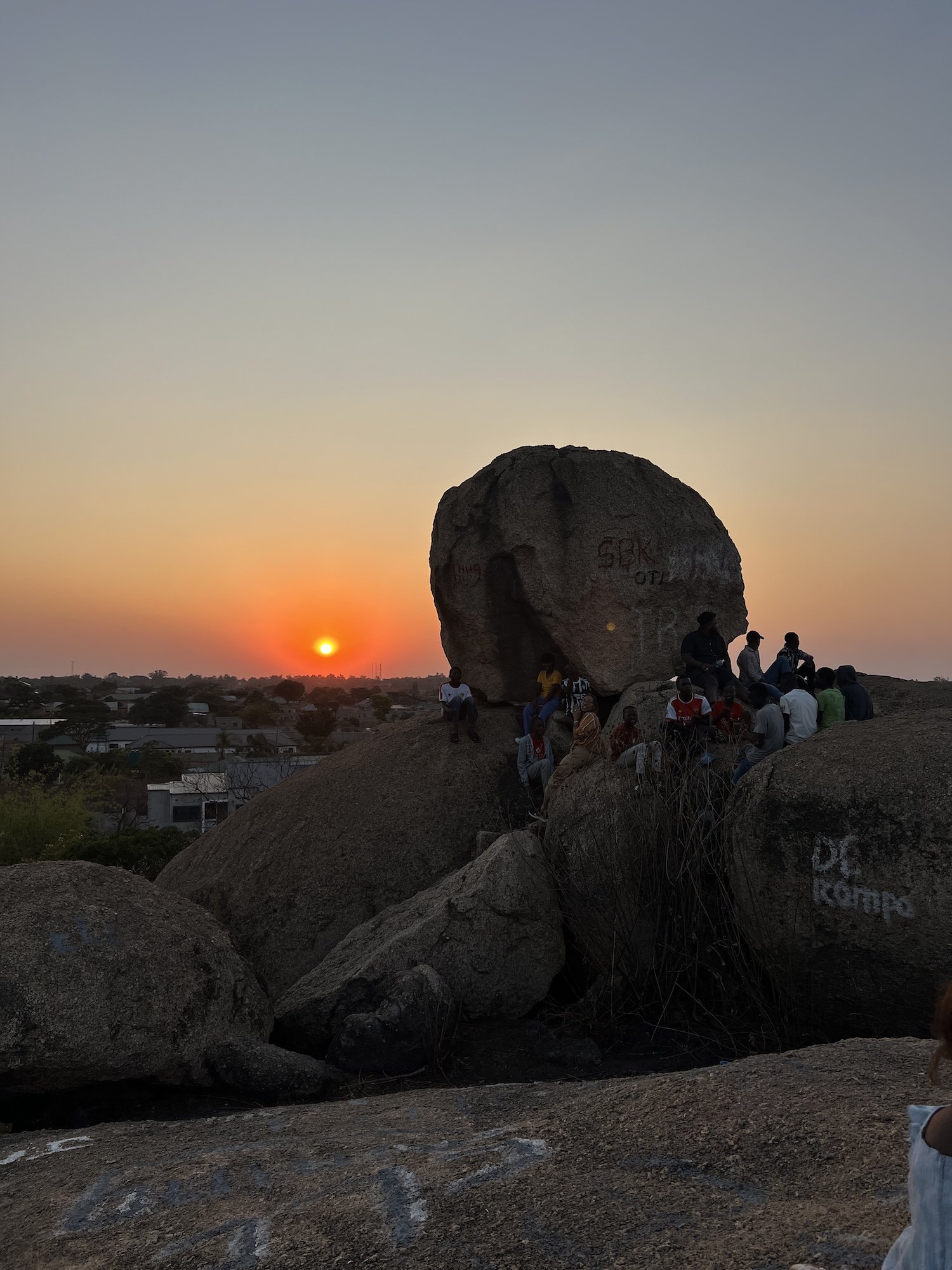 People gathering around large boulders as the sun sets in the background.