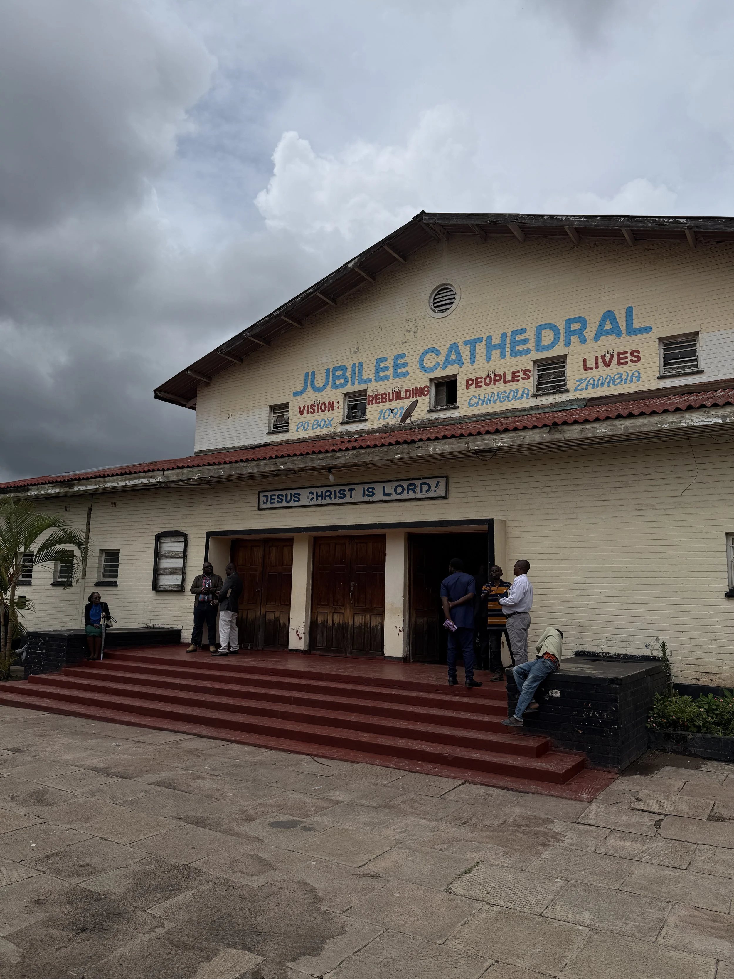 People standing on the steps of a church building named Jubilee Cathedral, with a sign reading 'Jesus Christ is Lord' above the entrance. The building has a sign about its vision of rebuilding lives.