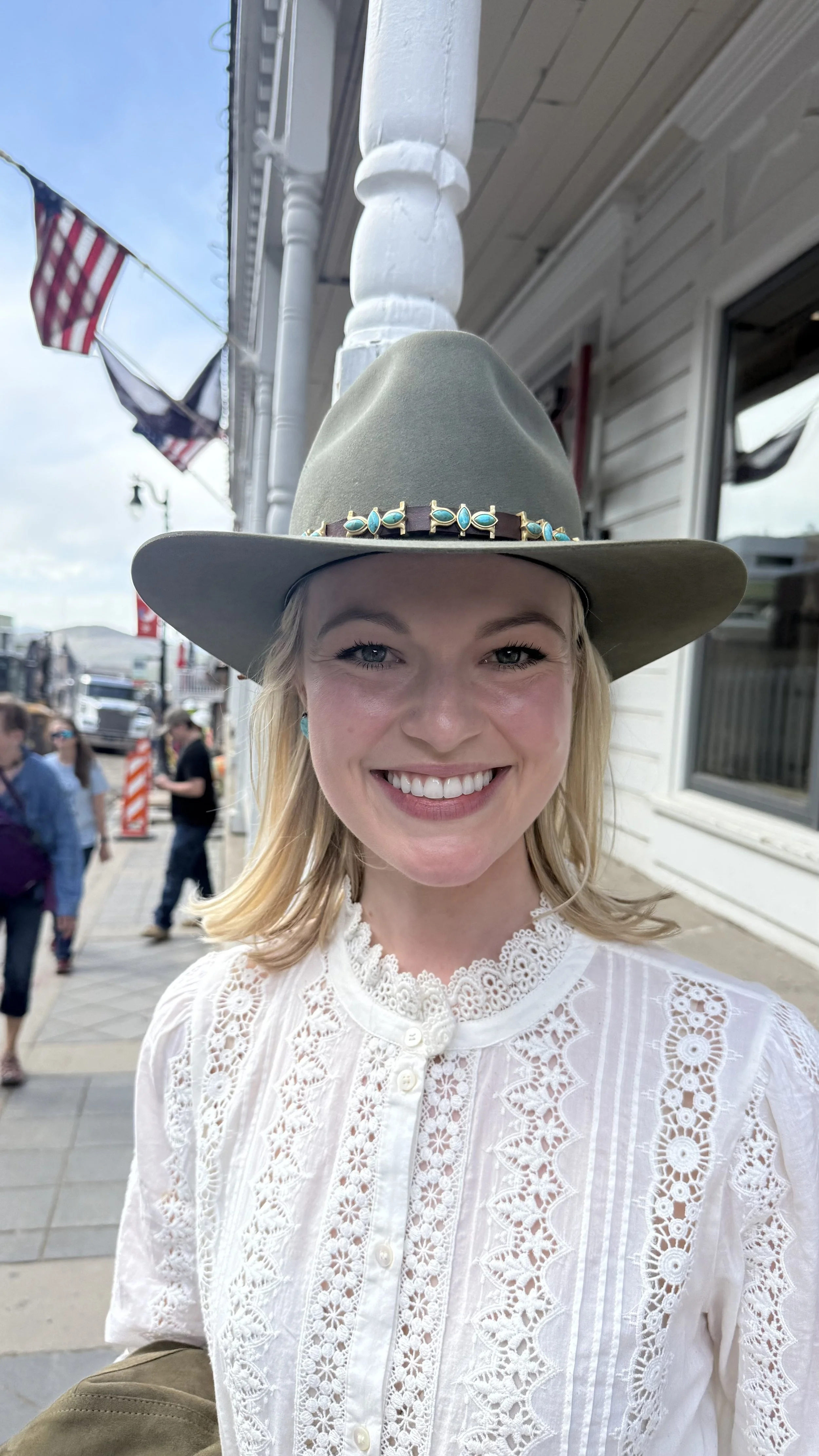 A smiling woman with blonde hair wearing a large gray wide-brimmed hat with turquoise accents and a white embroidered blouse, standing on a busy street with people and American flags in the background.