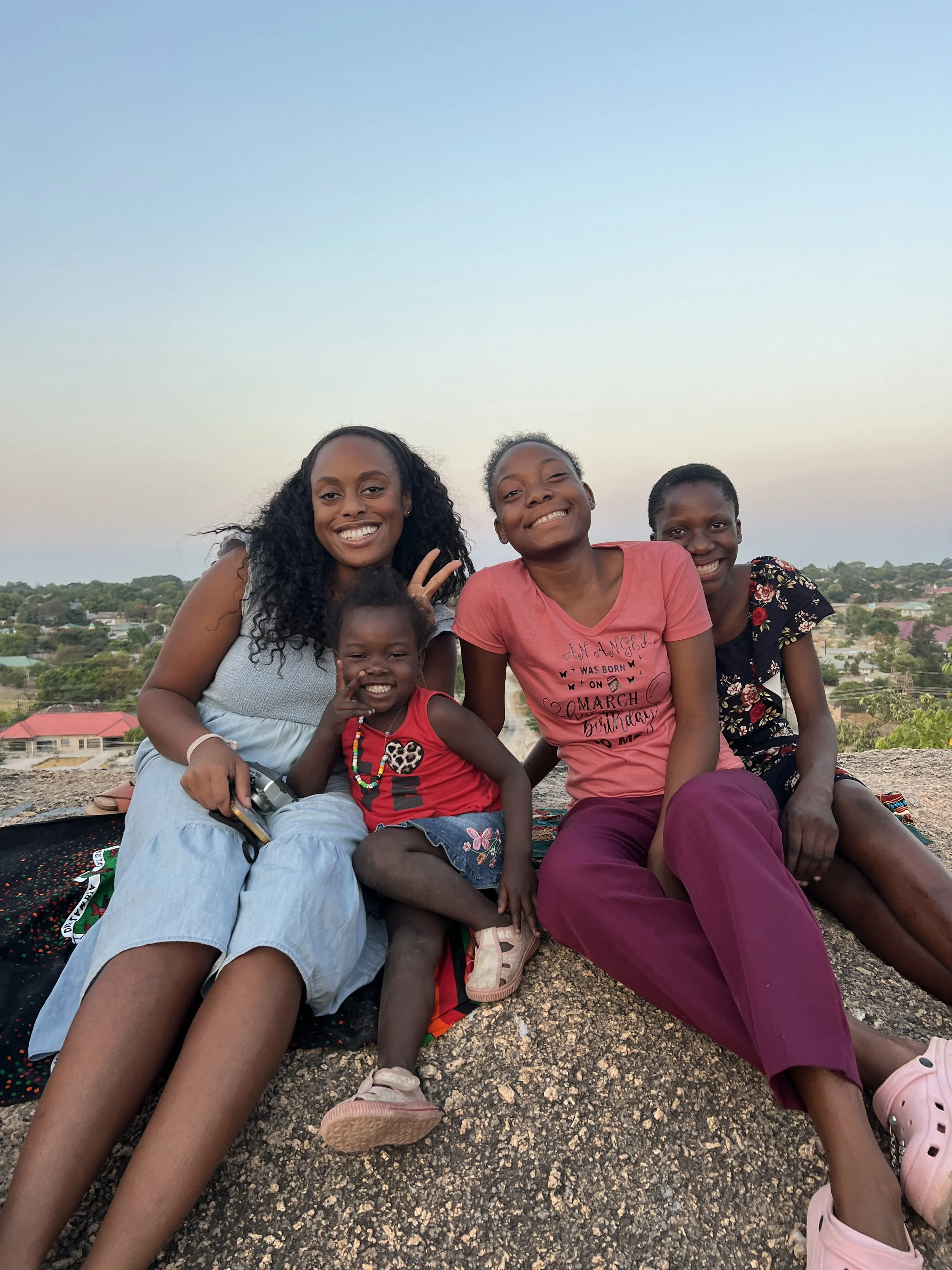 Group of four girls sitting on a rocky surface outdoors, smiling at the camera, with a distant landscape and houses in the background during sunset.