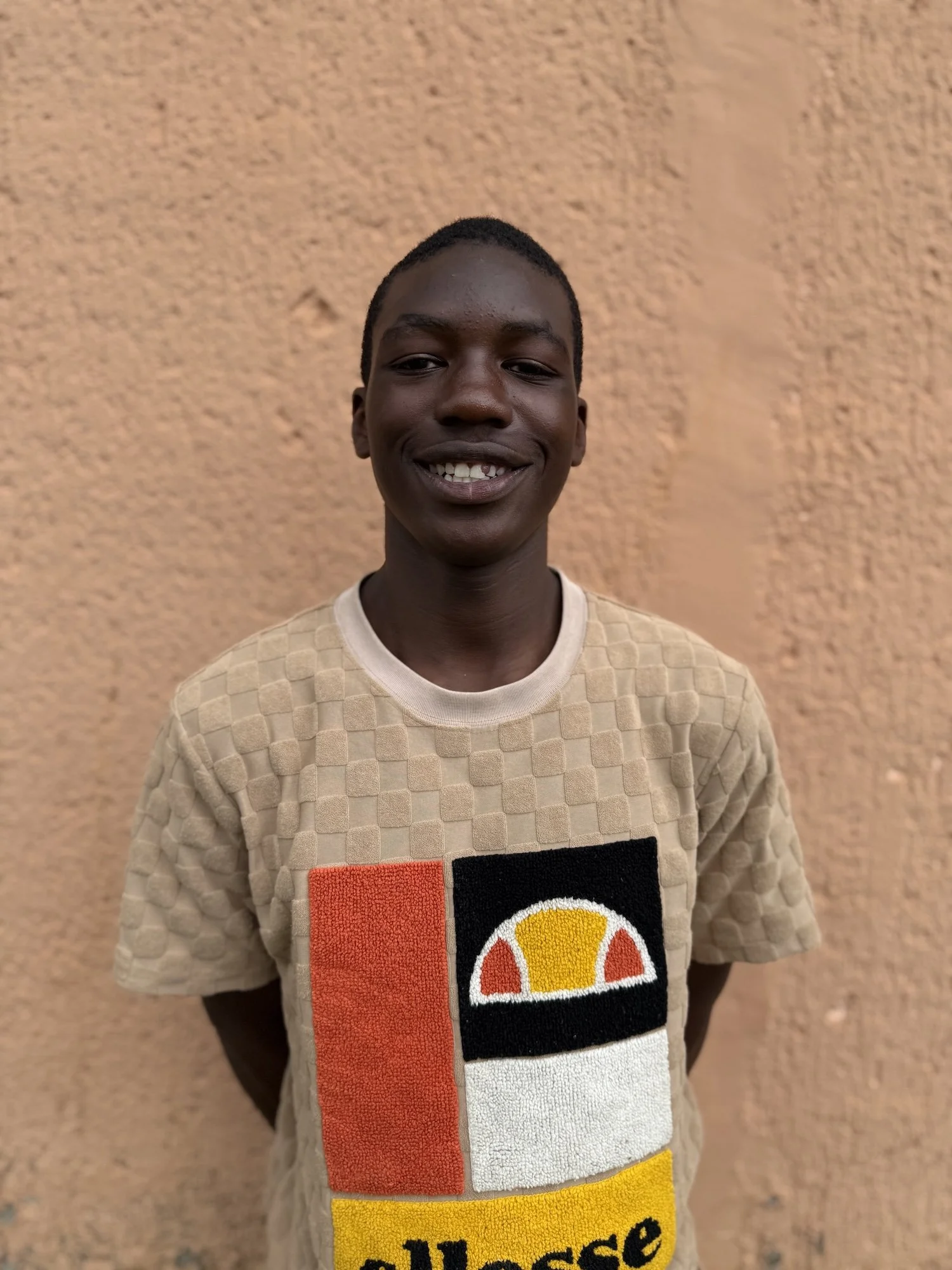 A young man standing against a textured beige wall, smiling and wearing a beige shirt with a colorful logo.