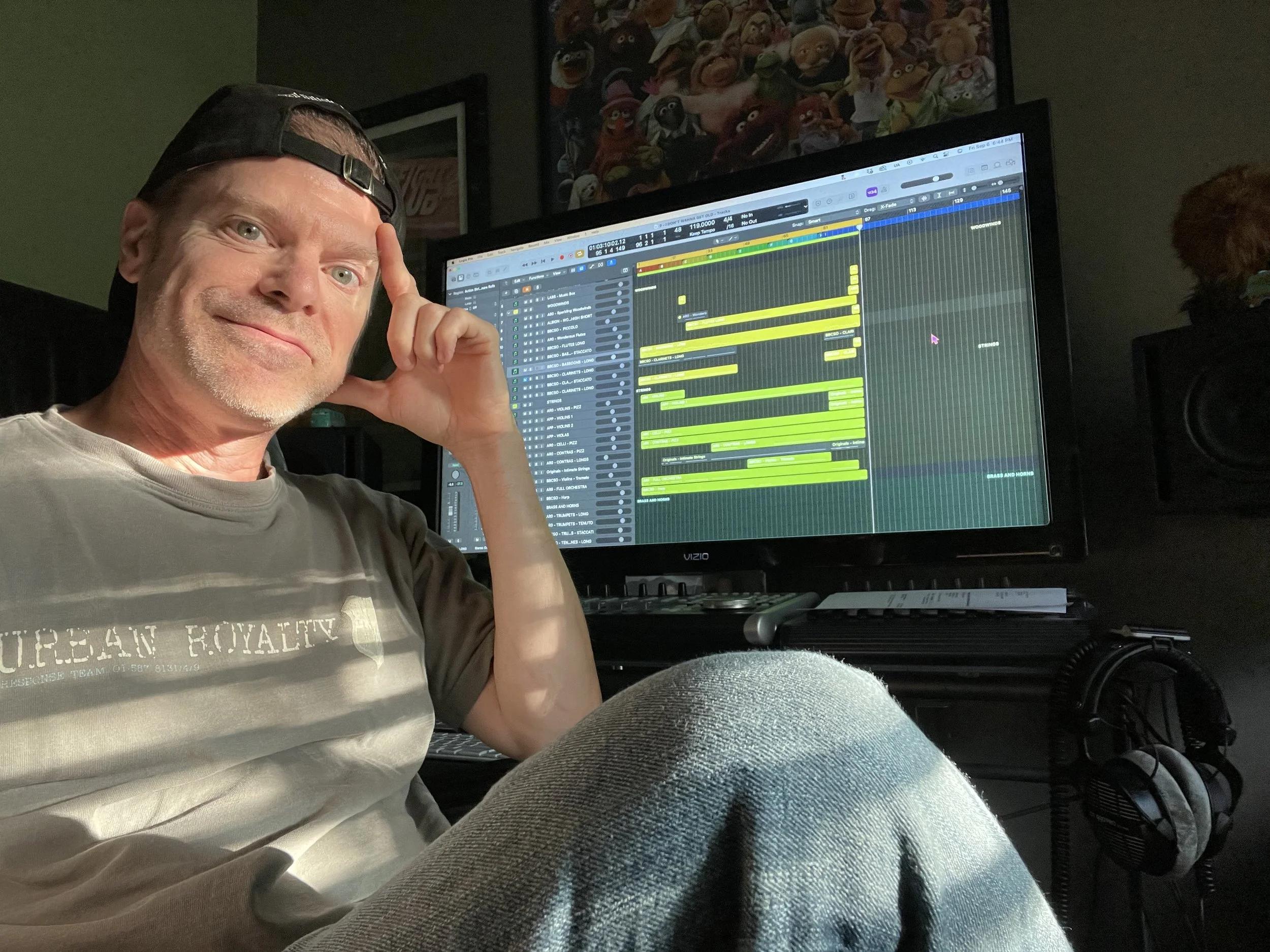 Smiling man with a backwards cap sitting in front of a computer screen displaying music production software, in a recording studio with posters on the wall and headphones hanging on the side.