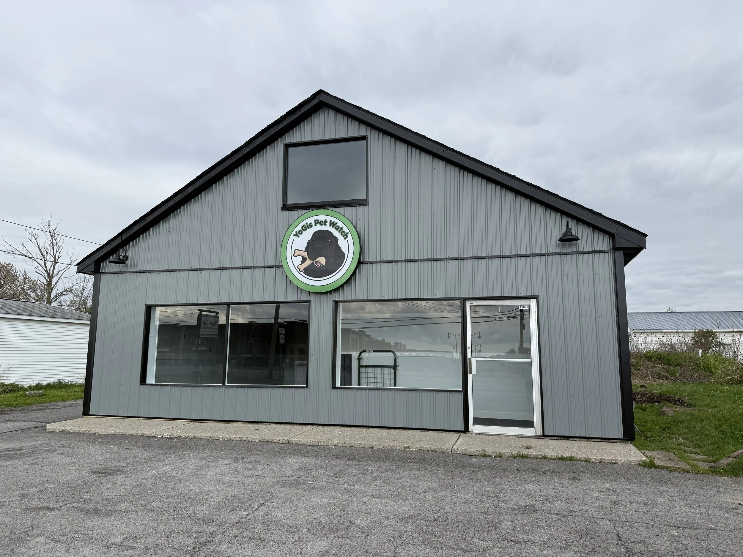A gray commercial building with a large window on the upper floor and a sign featuring a black dog holding a bone, indicating it is a pet watch business. The sky is cloudy and there are neighboring buildings and trees visible.