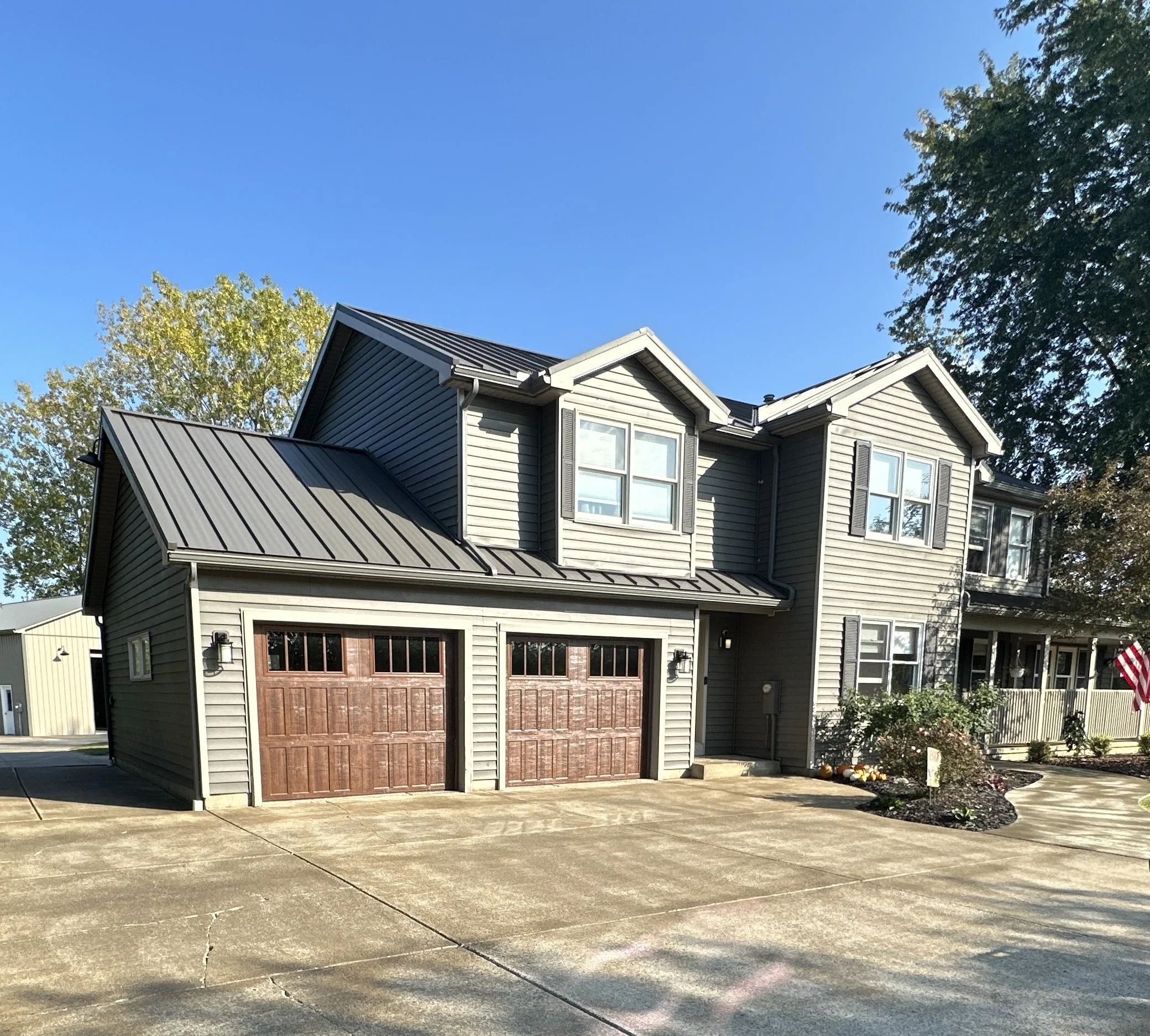A gray house with a gabled metal roof, large windows, and a wooden garage door, situated in a suburban neighborhood with trees and a concrete driveway.