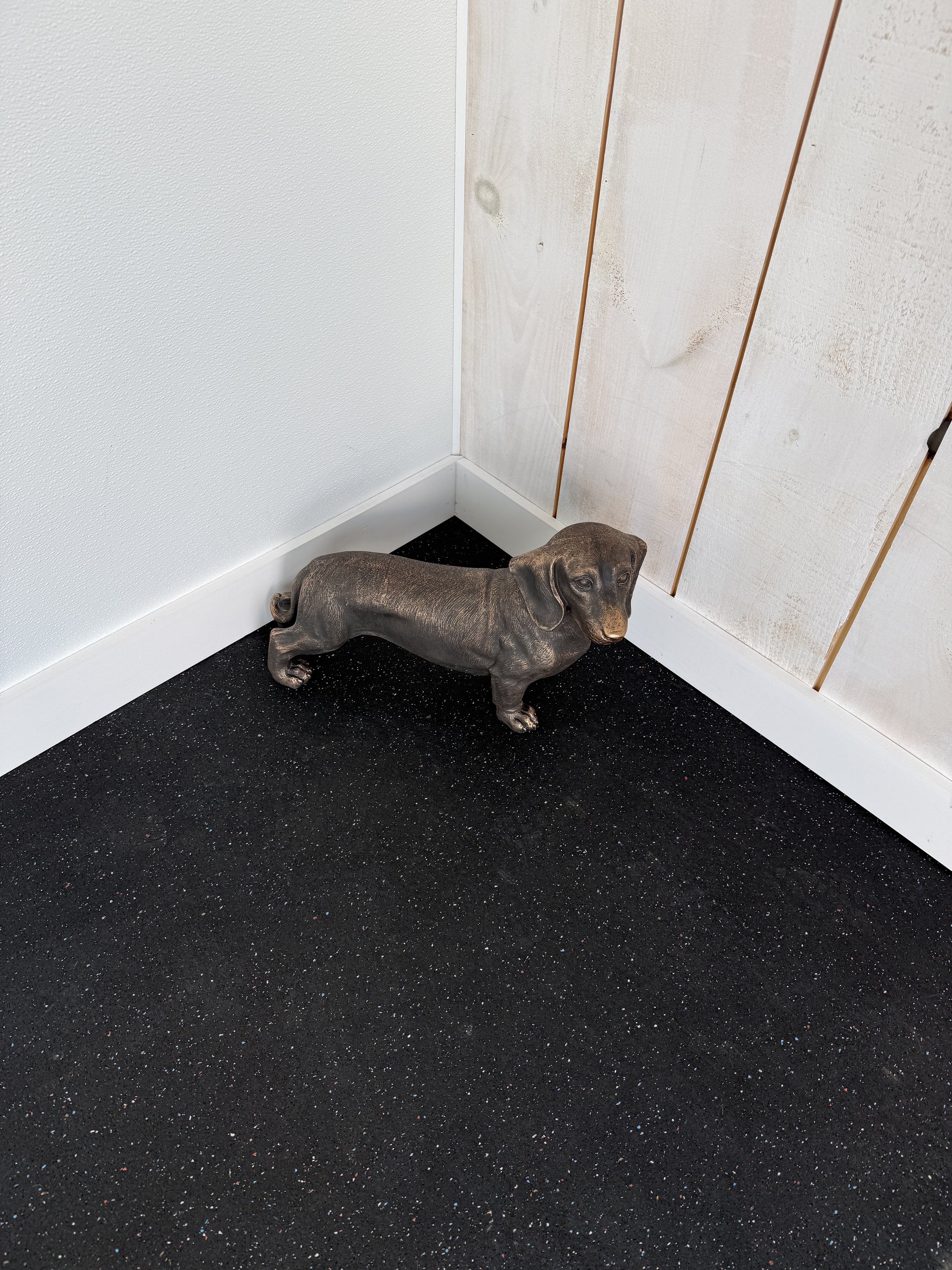 A gray statue of a dachshund dog positioned on a black speckled carpet, against white wall and paneling.