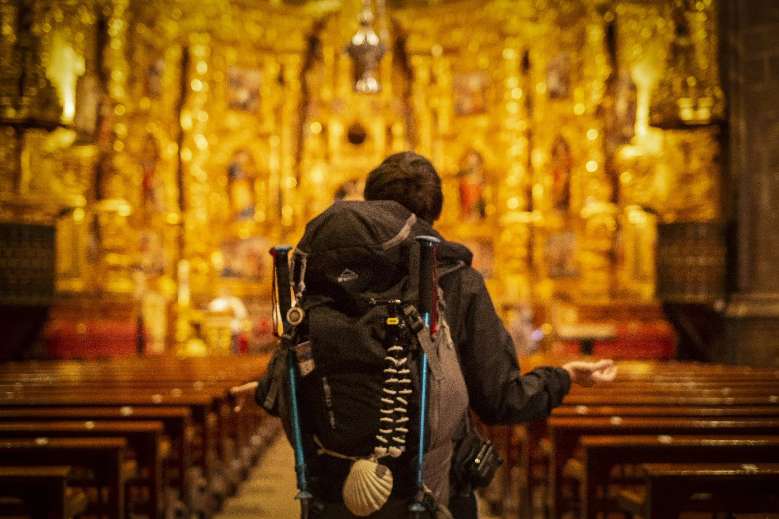 A person with short hair, wearing a backpack, standing in a warmly lit church interior with gold ornate decorations on the wall.