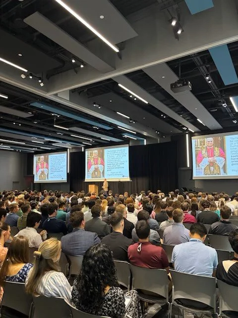 A large conference room filled with a diverse audience attending a presentation. The room has multiple large screens displaying slides, with a speaker at a podium. The ceiling features modern lighting and acoustic panels.