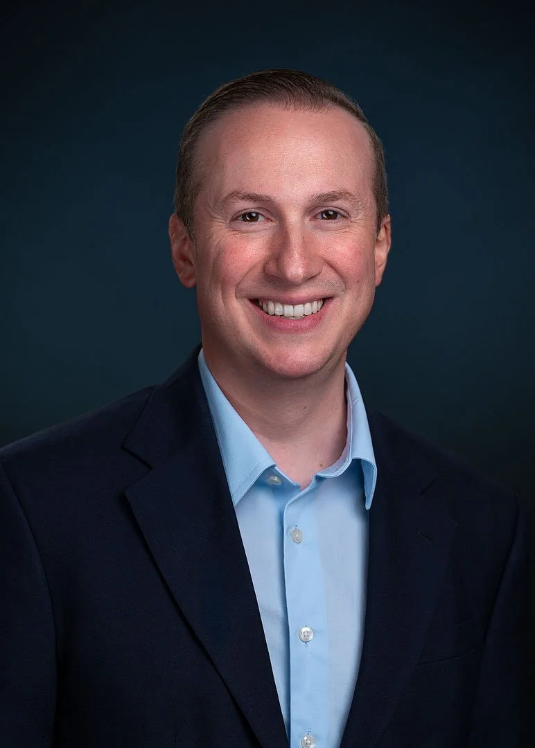 Headshot of a smiling man in a dark suit jacket and light blue collared shirt, against a dark background.