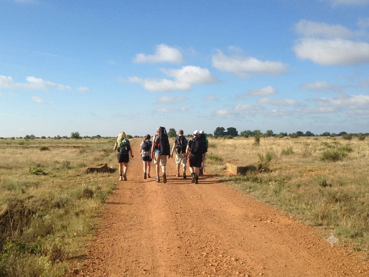 Group of six hikers walking on a dirt trail through a flat, open grassland under a blue sky with scattered clouds.