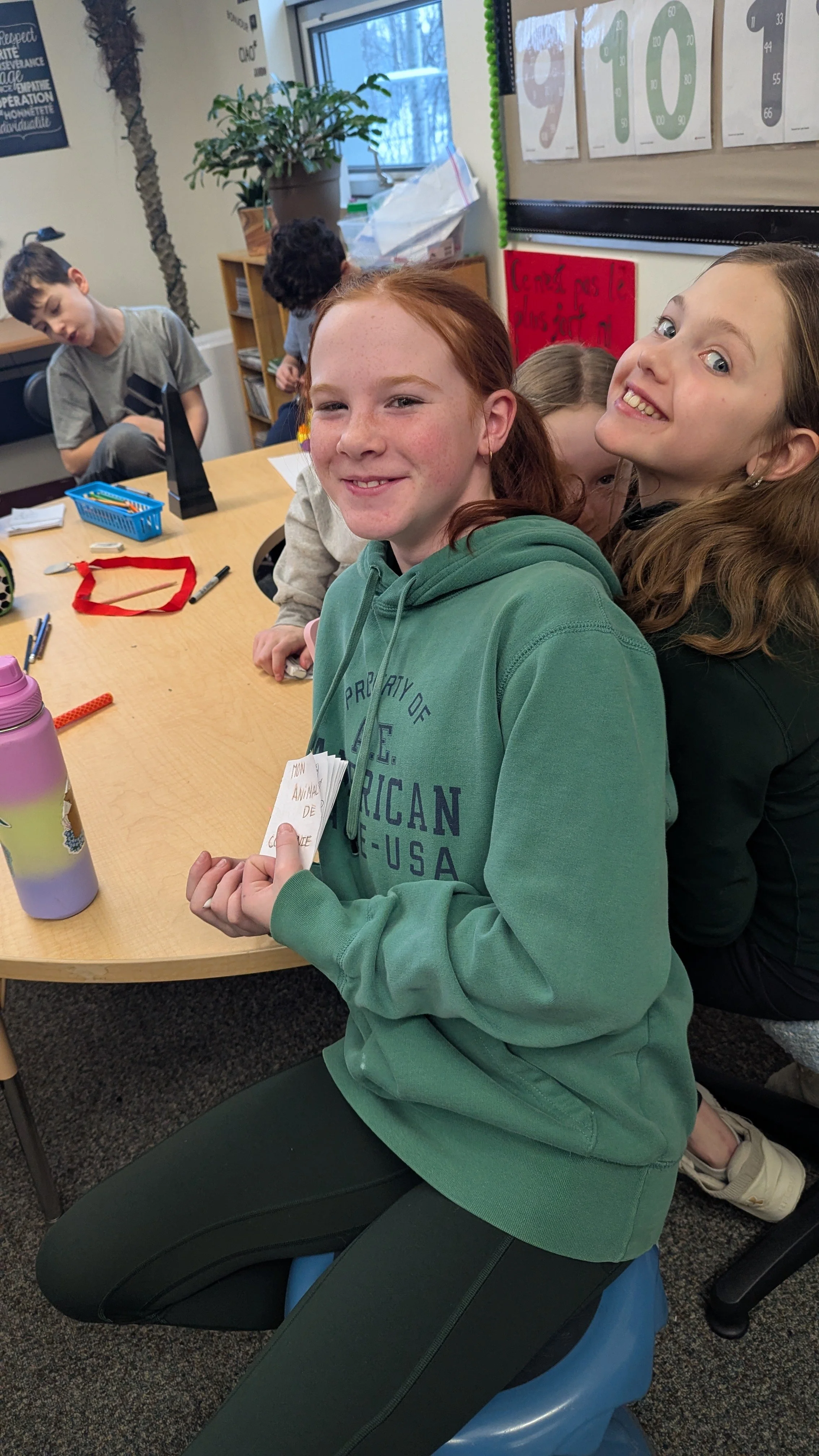 Une jeune fille assise sur un ballon bleu dans une classe, entourée d'autres enfants, tenant des papiers, avec un tableau de chiffres en arrière-plan.