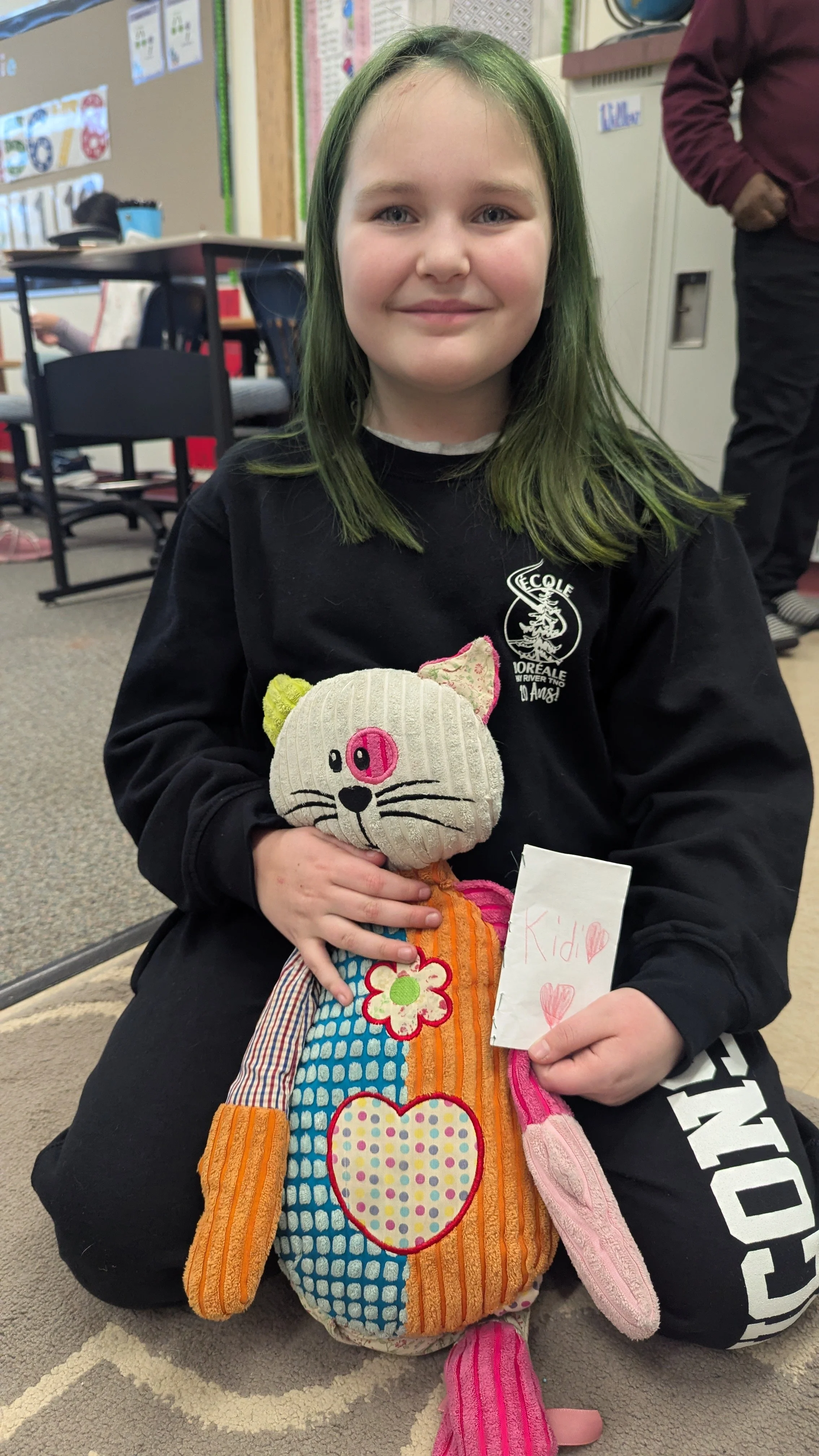 Une jeune fille souriante tenant une poupée en tissu colorée et un petit papier avec le mot 'Kid' et des cœurs rédigés à la main, assise sur un tapis dans une salle de classe.