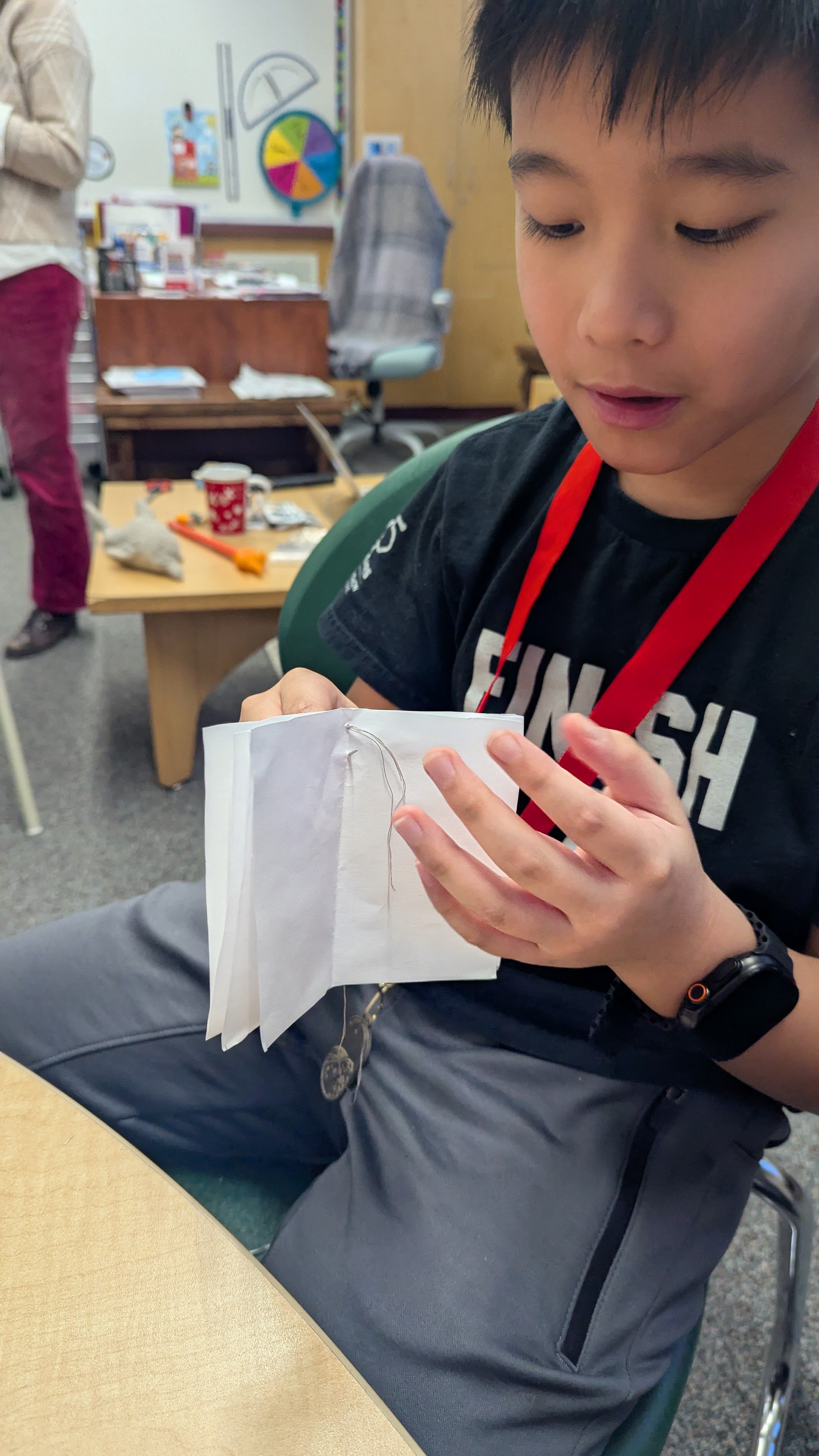 Jeune garçon à lunettes assis, regardant un petit livre ou carnet blanc avec fil. Autour de lui, une salle avec un bureau, une chaise, des objets divers et un tableau blanc avec un cercle coloré.