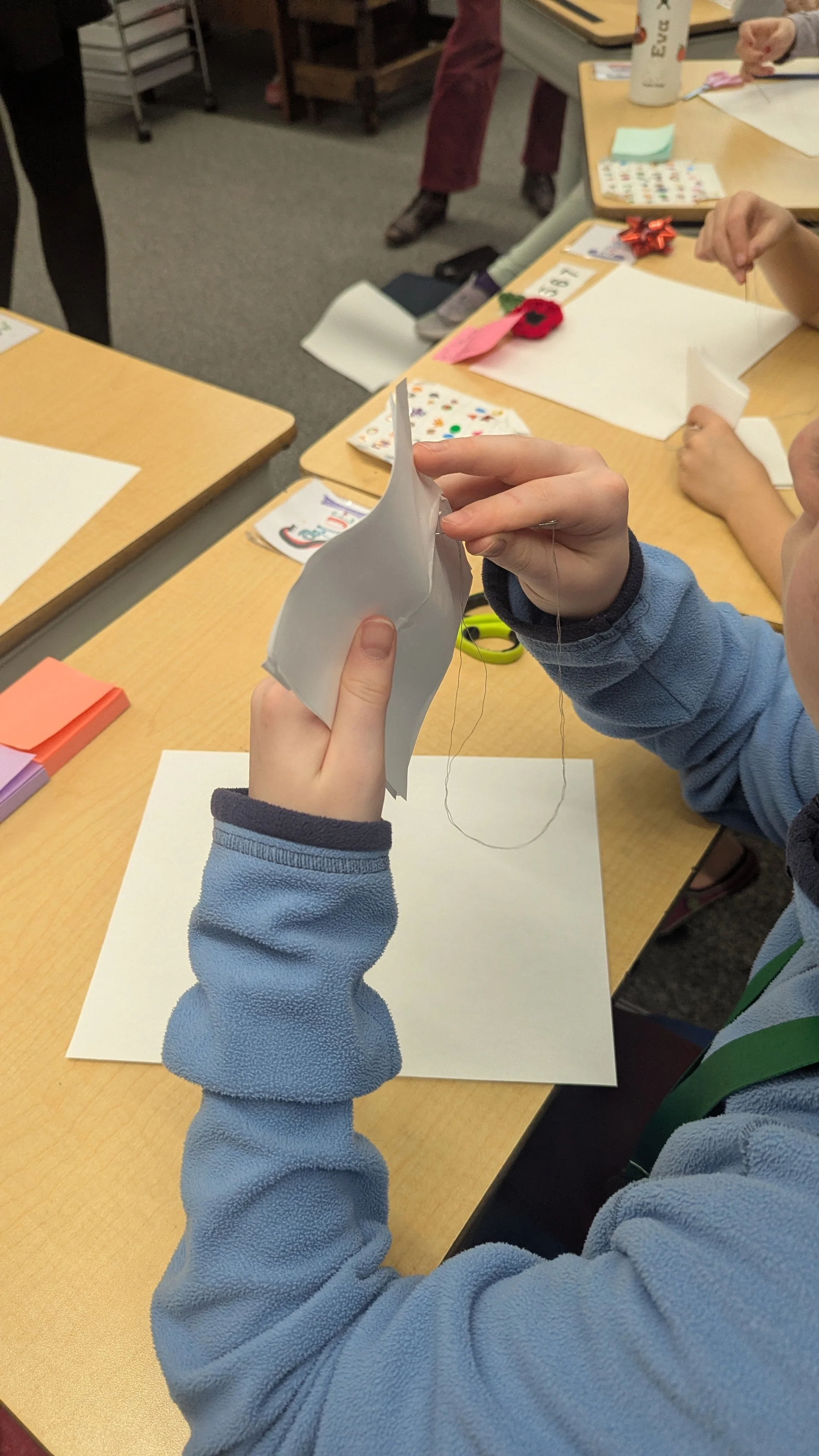 Enfant en pull bleu tenant une feuille de papier pliée et une aiguille à coudre dans une école, avec d'autres enfants et des fournitures scolaires sur la table.