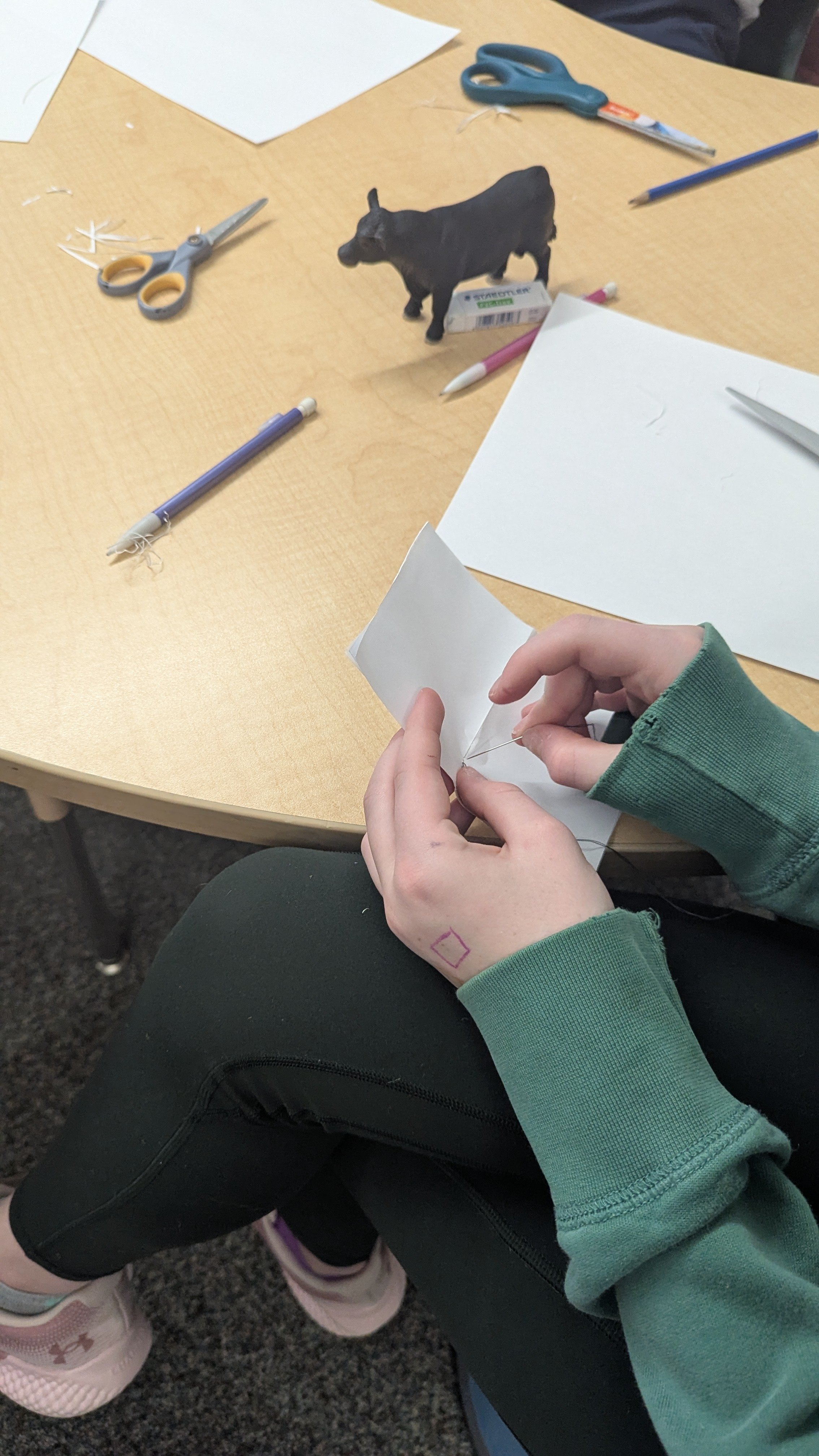 Une personne en train de faire de la couture ou du bricolage, utilisant une aiguille sur un morceau de tissu blanc, avec des ciseaux, des stylos, et une figurine de vache sur la table.