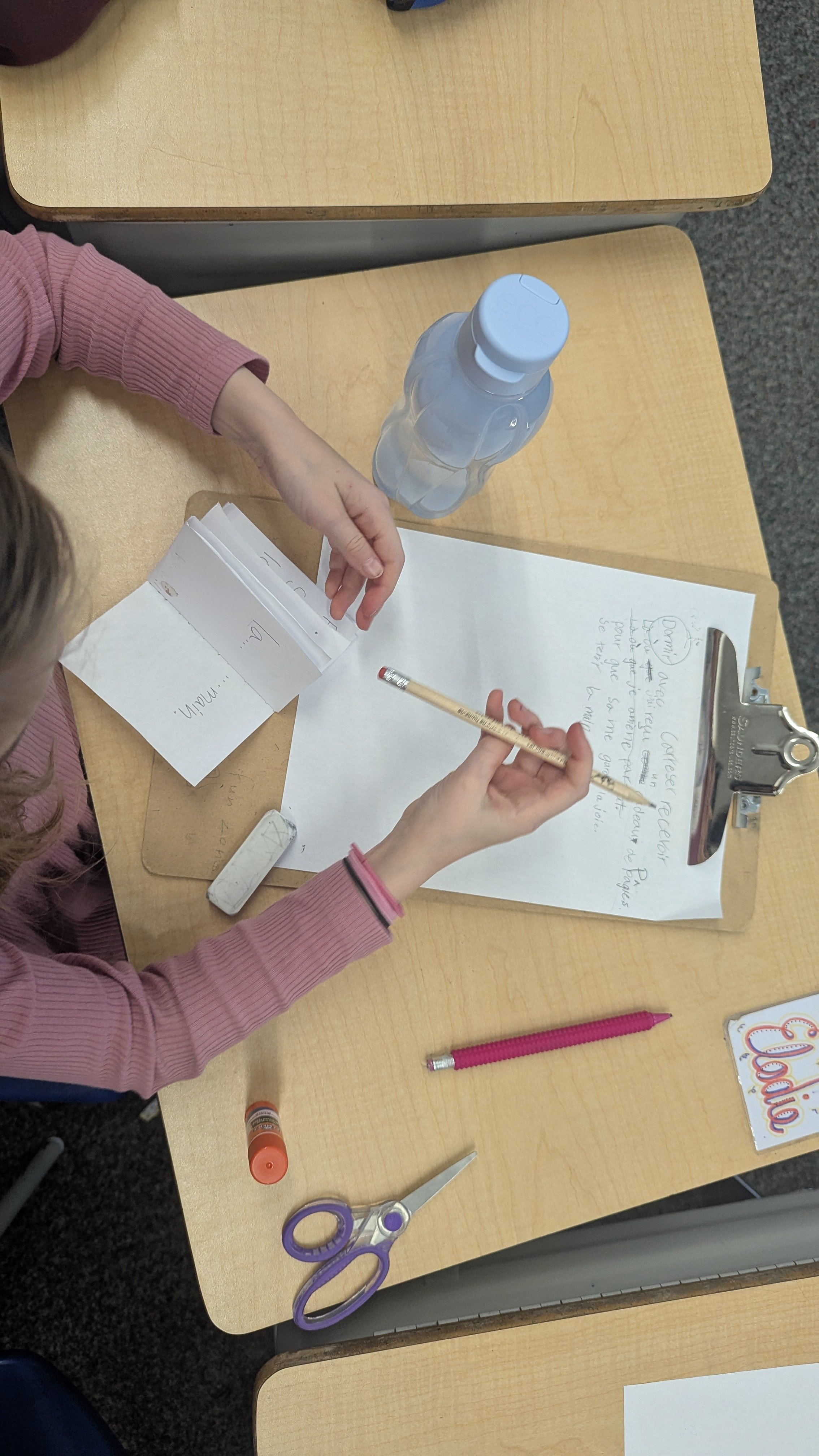 Une enfant écrit sur une feuille de papier à l'aide d'un crayon. Sur la table, il y a des ciseaux, un stylo, un tube de colle, une gomme, une bouteille d'eau, un ensemble de fiches et une clip.