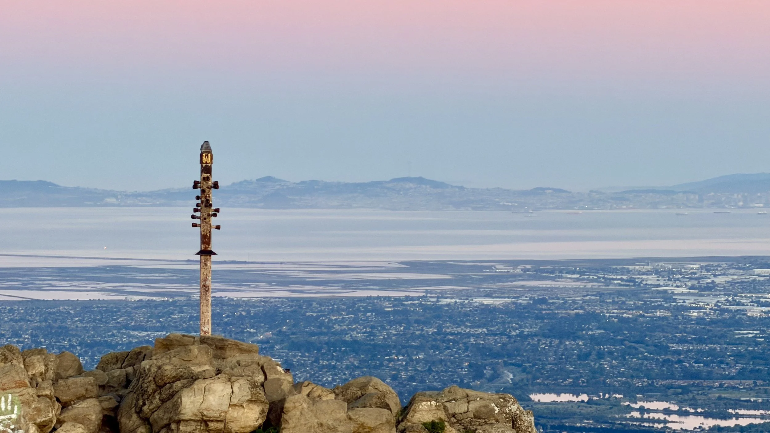 A weathered metal pole with radar and antennas stands on a rocky elevation overlooking a snow-covered valley with mountains in the distance under a pastel pink, blue, and purple sky.