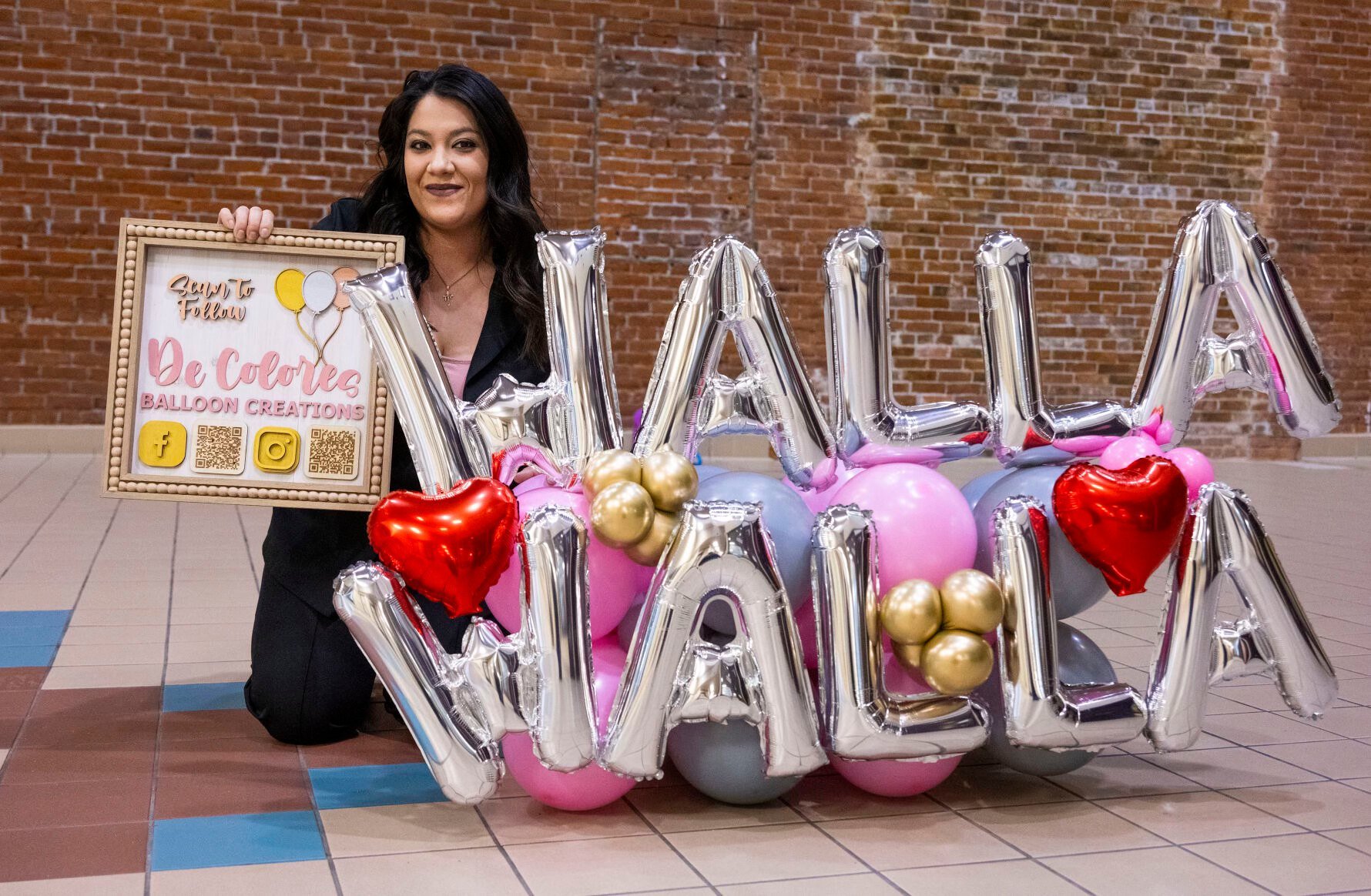 A woman holding a sign and kneeling next to a colorful balloon display with the name 'De Coates' and the phrase 'Say to Follow' on it. The balloon arrangement spells out 'WAL' with large balloons and includes pink, gold, black, and gray balloons, as well as foil heart-shaped balloons.
