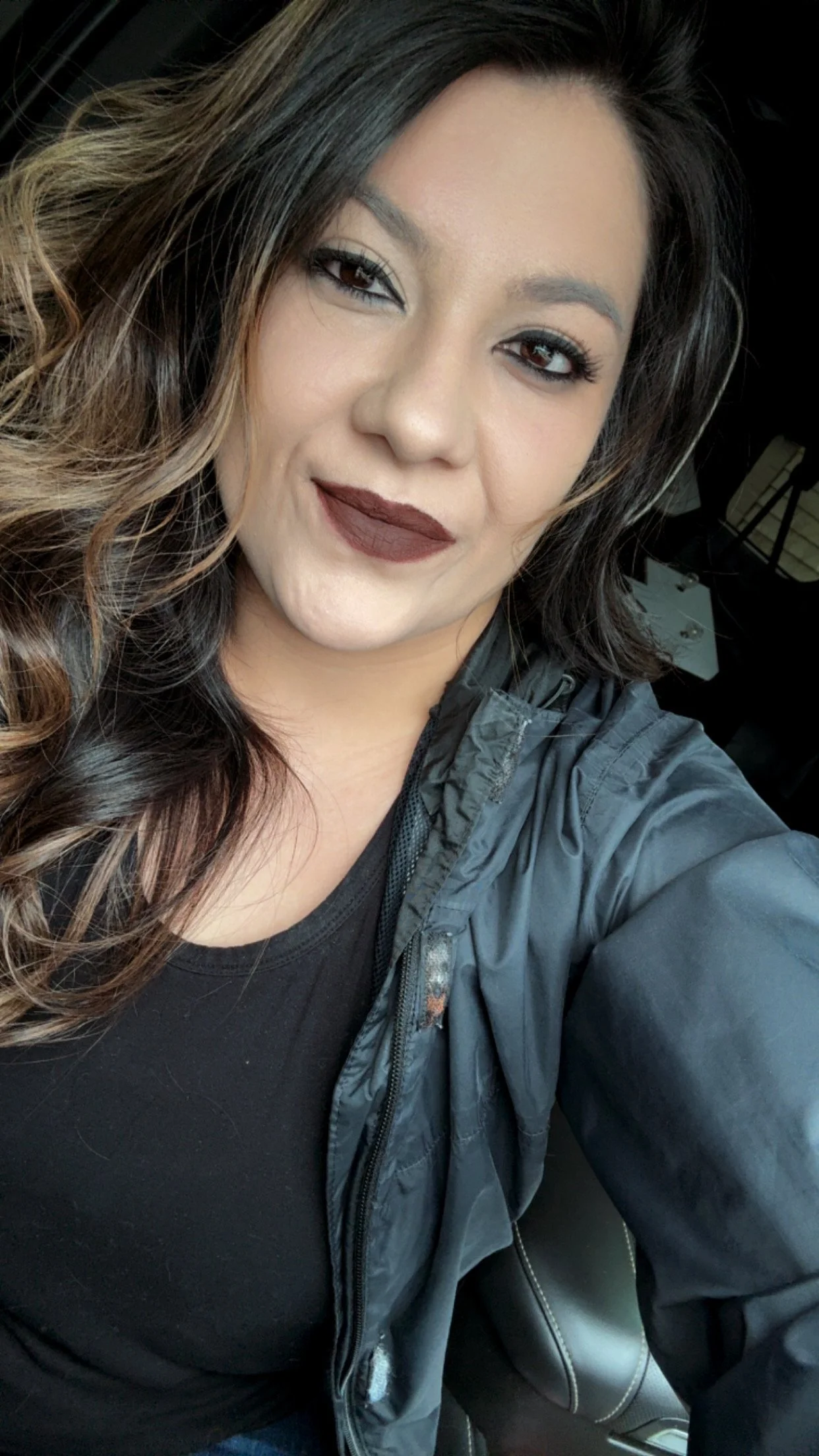 A woman with wavy hair, wearing makeup with dark lipstick and eyeshadow, taking a selfie inside a car. She is dressed in a black top and a dark jacket.