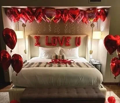 Decorated bedroom with red heart-shaped balloons, love-themed pillows, and a banner that reads 'I LOVE U' for a romantic occasion.