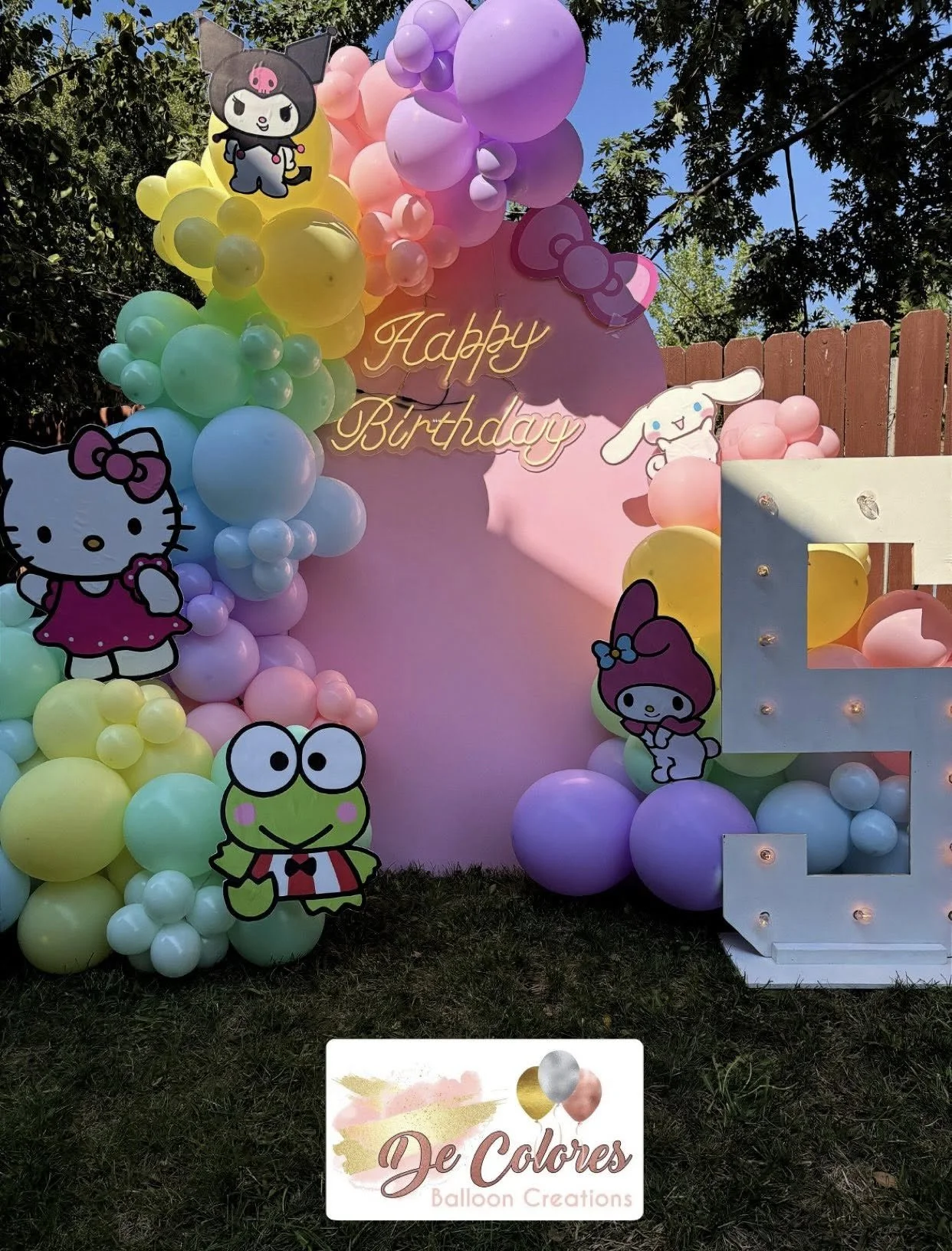 Colorful balloon arrangement with Hello Kitty characters and a pink backdrop that says 'Happy Birthday,' decorated with balloons and lights at an outdoor birthday celebration.
