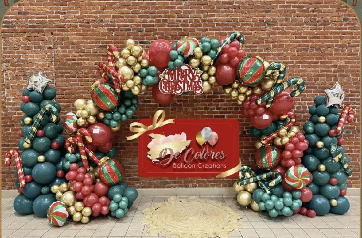 Christmas balloon arch with red, green, gold, and plaid balloons, stars, and peppermint candy decorations in front of a brick wall, with a red sign reading "De Colores Balloon Creations".