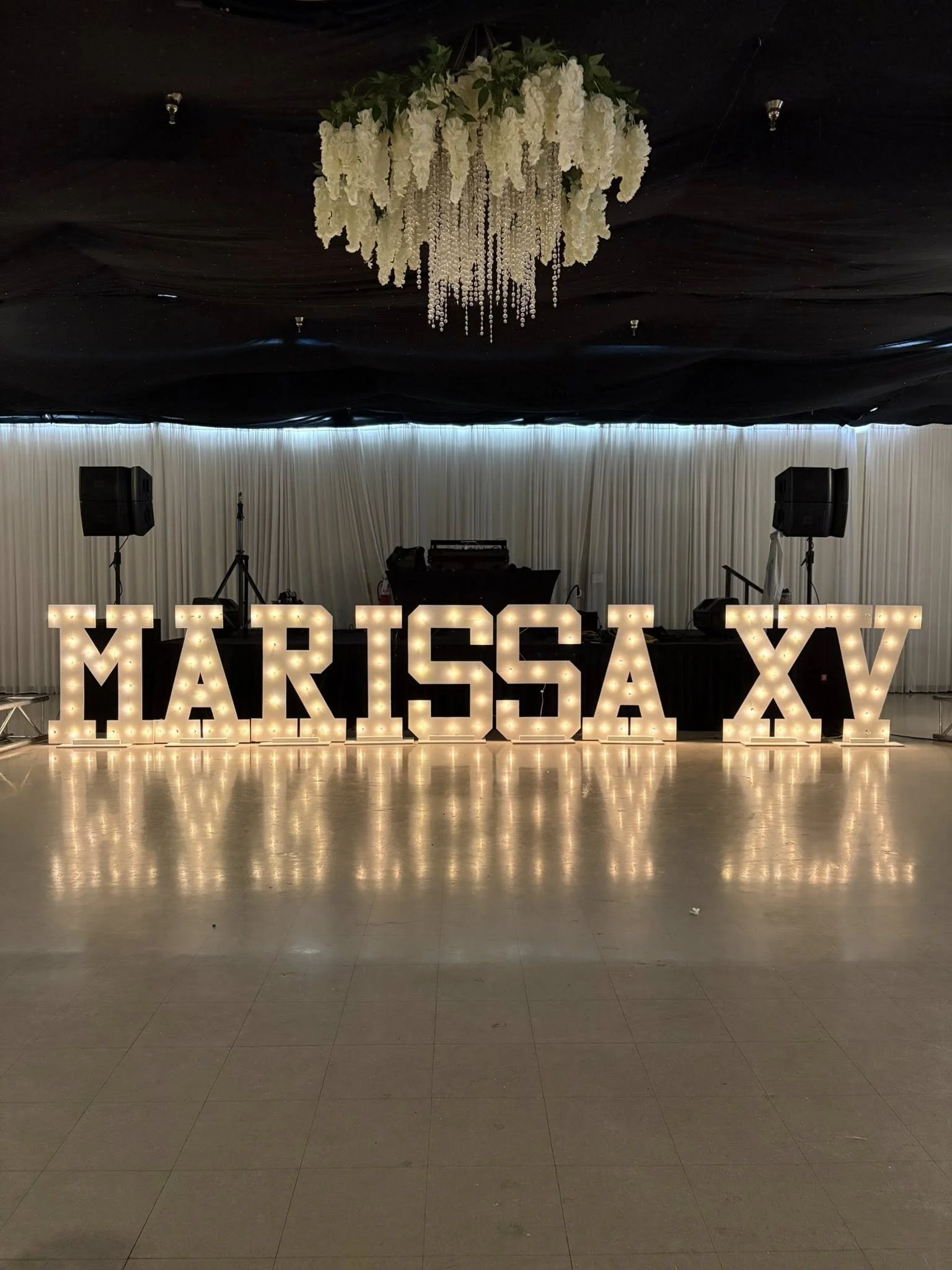 Decorative wedding or event setup with large illuminated letters spelling 'MARTINA XY', a chandelier with white flowers hanging from a black ceiling, and a DJ station with speakers and lighting equipment, all on a reflective floor.