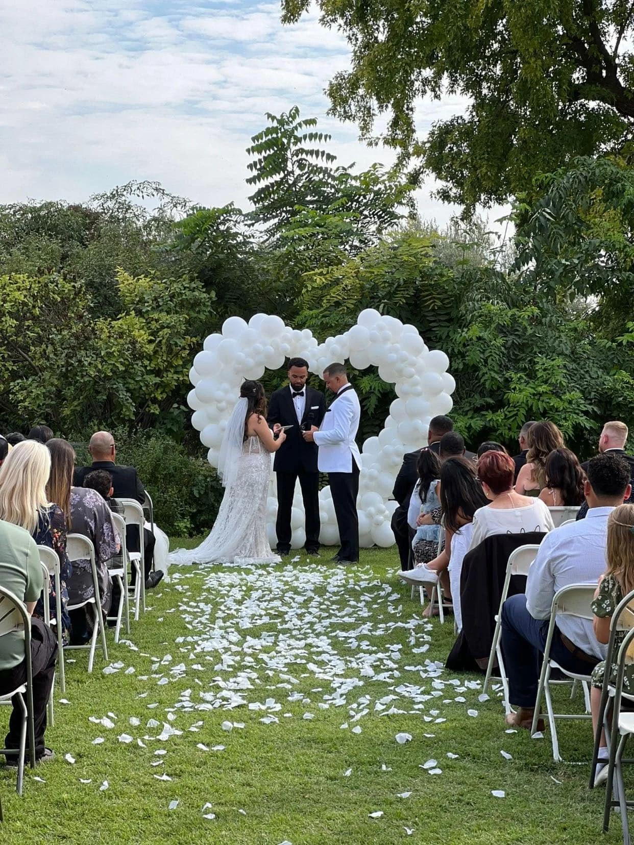 An outdoor wedding ceremony with a bride and groom standing before an officiant, in front of a heart-shaped balloon arch. Guests are seated on either side, and there are white flower petals on the grass aisle.