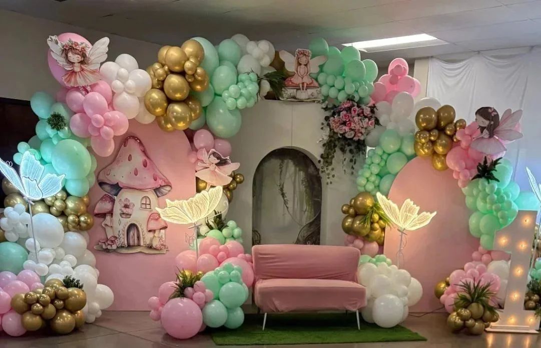 Decorative backdrop with pastel pink, mint green, white, and gold balloons, fairy-themed cutouts, and illuminated butterfly decorations, featuring a pink loveseat in front.