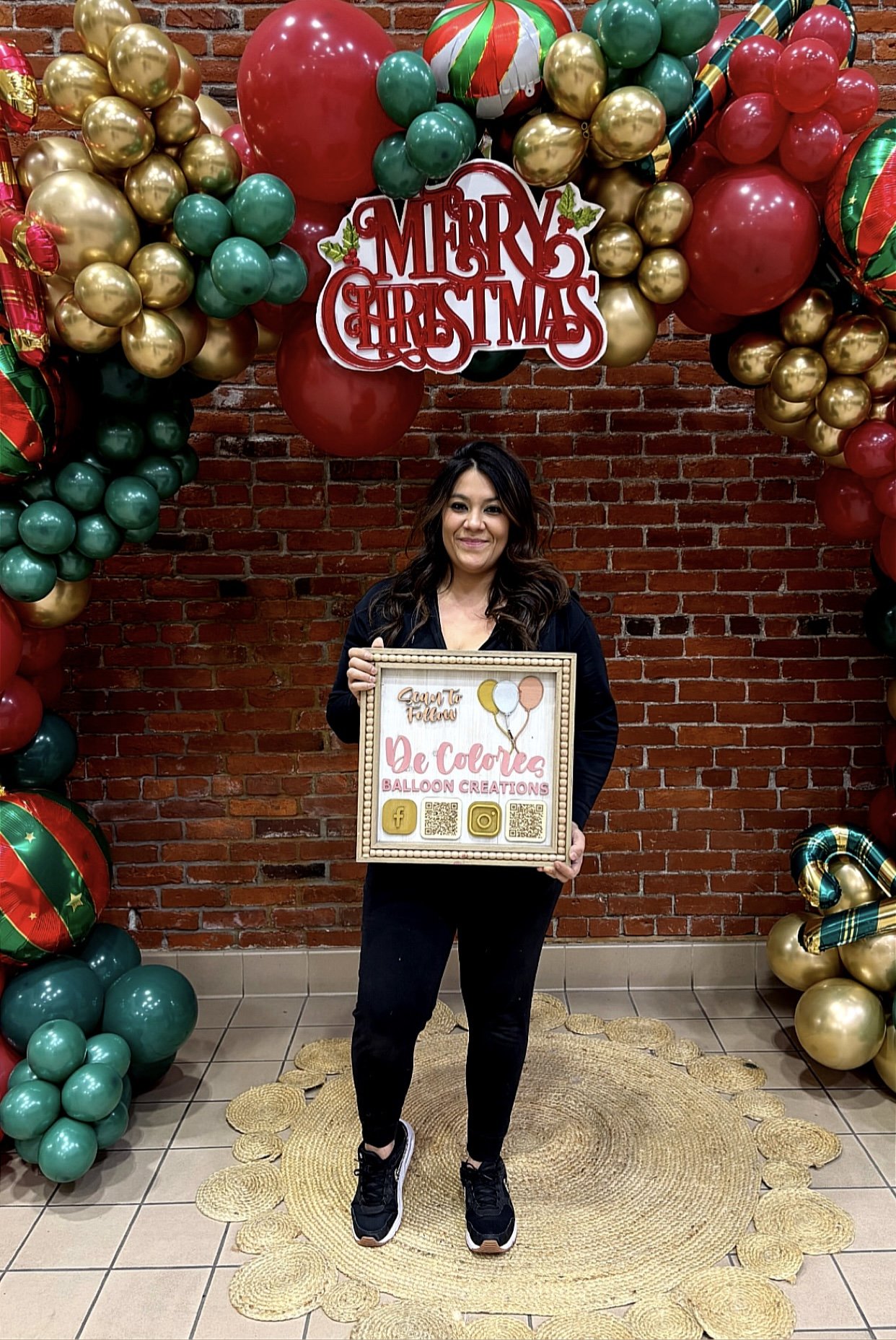 A woman standing on a yellow woven rug in front of a red brick wall decorated with Christmas balloons and a "Merry Christmas" sign. She is holding a framed sign with balloons, social media icons, and QR codes, and is smiling.