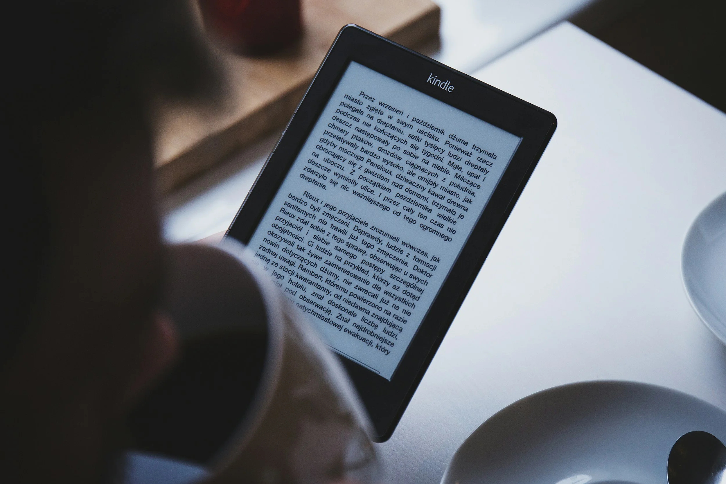A person holding a Kindle e-reader displaying a page with text in Polish. The scene is set on a white surface next to a white cup and a wooden object, with a dark and blurred background.