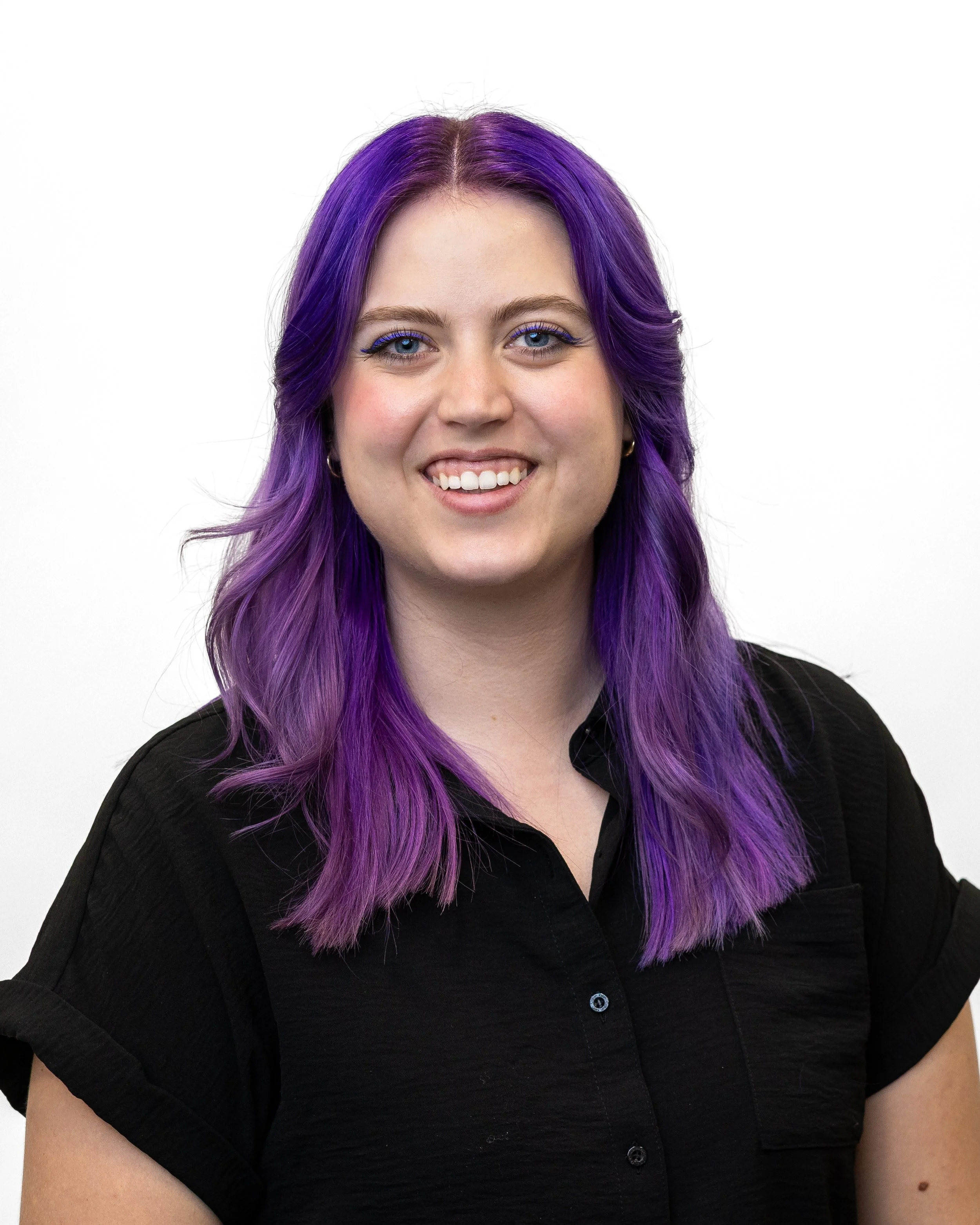 A woman with purple hair, wearing a black top, smiling, against a plain white background.