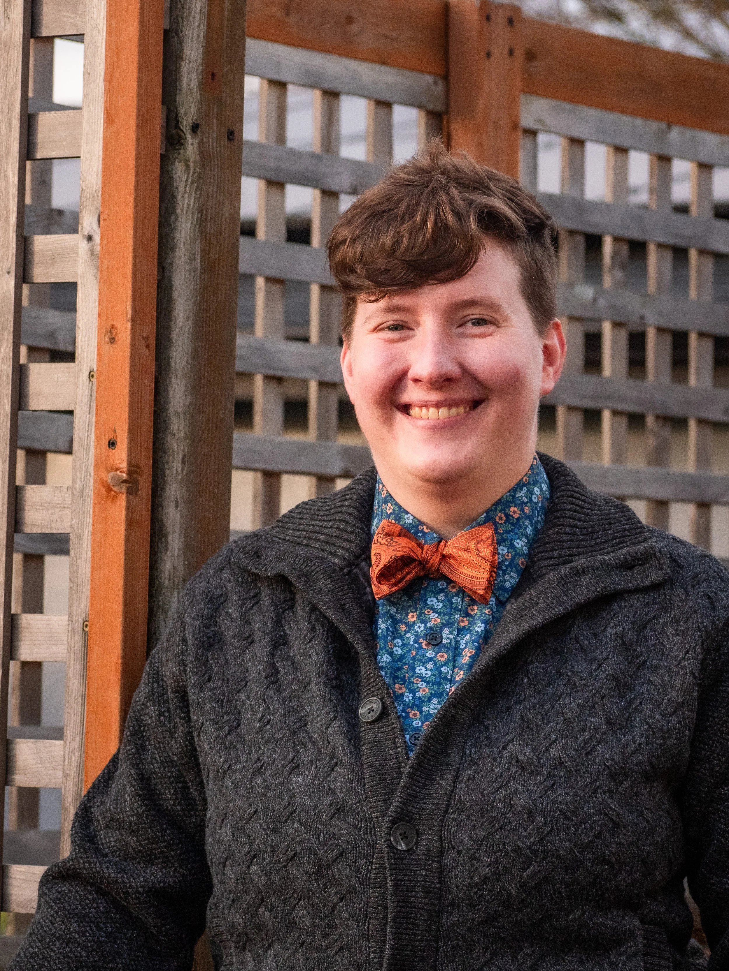 A young man with curly hair smiling outdoors, wearing a dark gray knit cardigan, a blue floral shirt, and an orange patterned bow tie, standing next to a wooden fence.