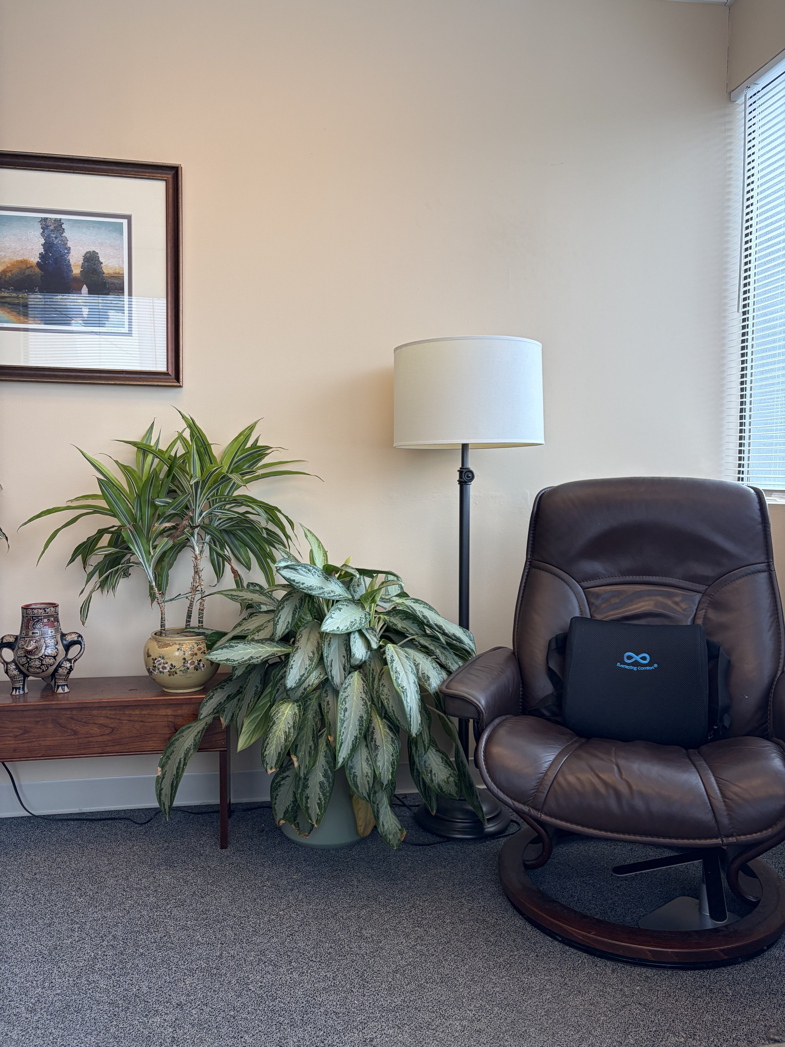 Office corner with a brown leather chair, a tall floor lamp, a wooden console table with two potted plants and a decorative vase, and a framed landscape painting on the wall.