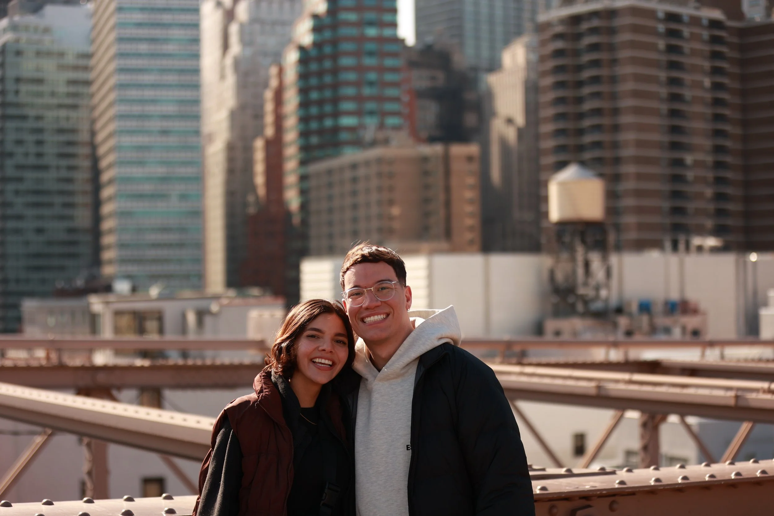 A young man and woman smiling together on a city rooftop with tall buildings in the background during daylight.