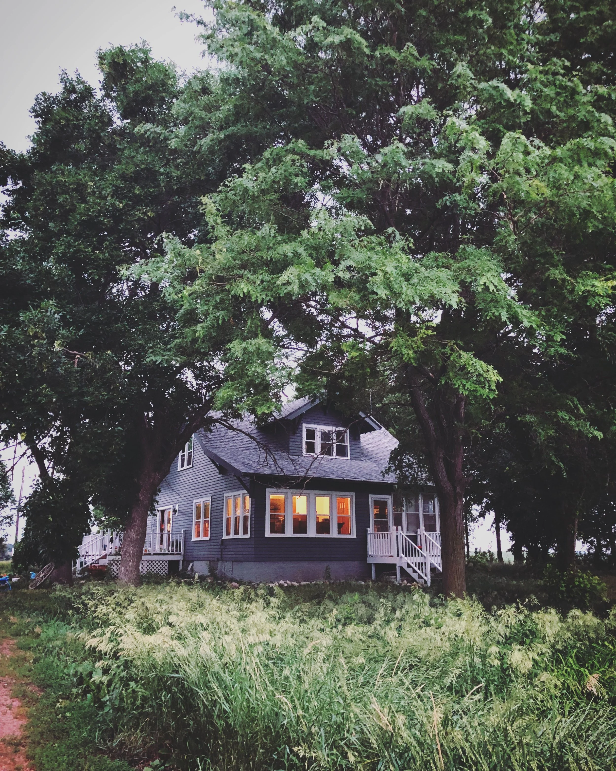 A house with a large front porch and multiple windows, surrounded by tall trees and lush green grass, during dusk with some windows illuminated.