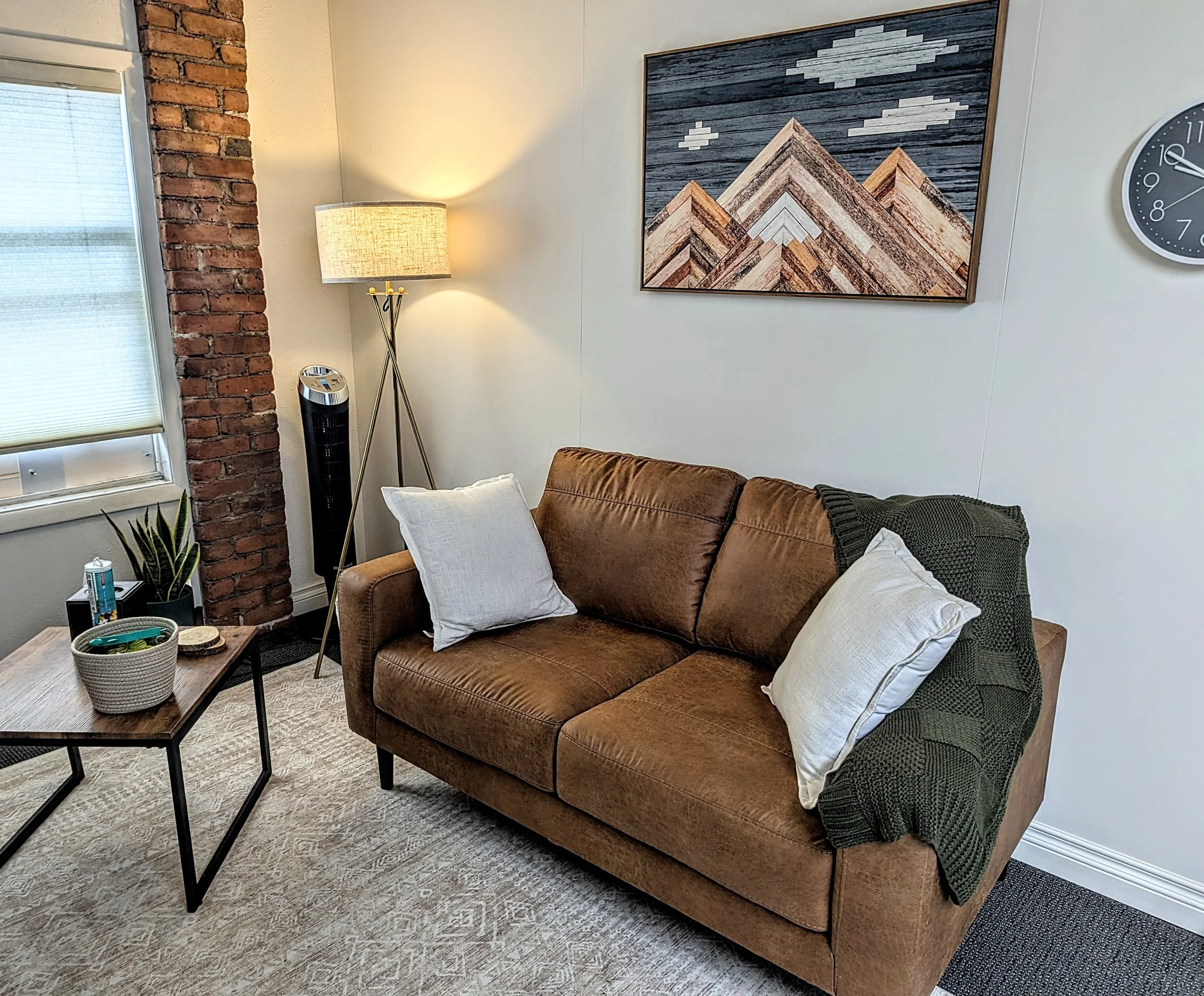 Living room with a brown leather sofa, throw pillows, a coffee table, a lamp, a brick wall, a window with blinds, a potted plant, and wall art.
