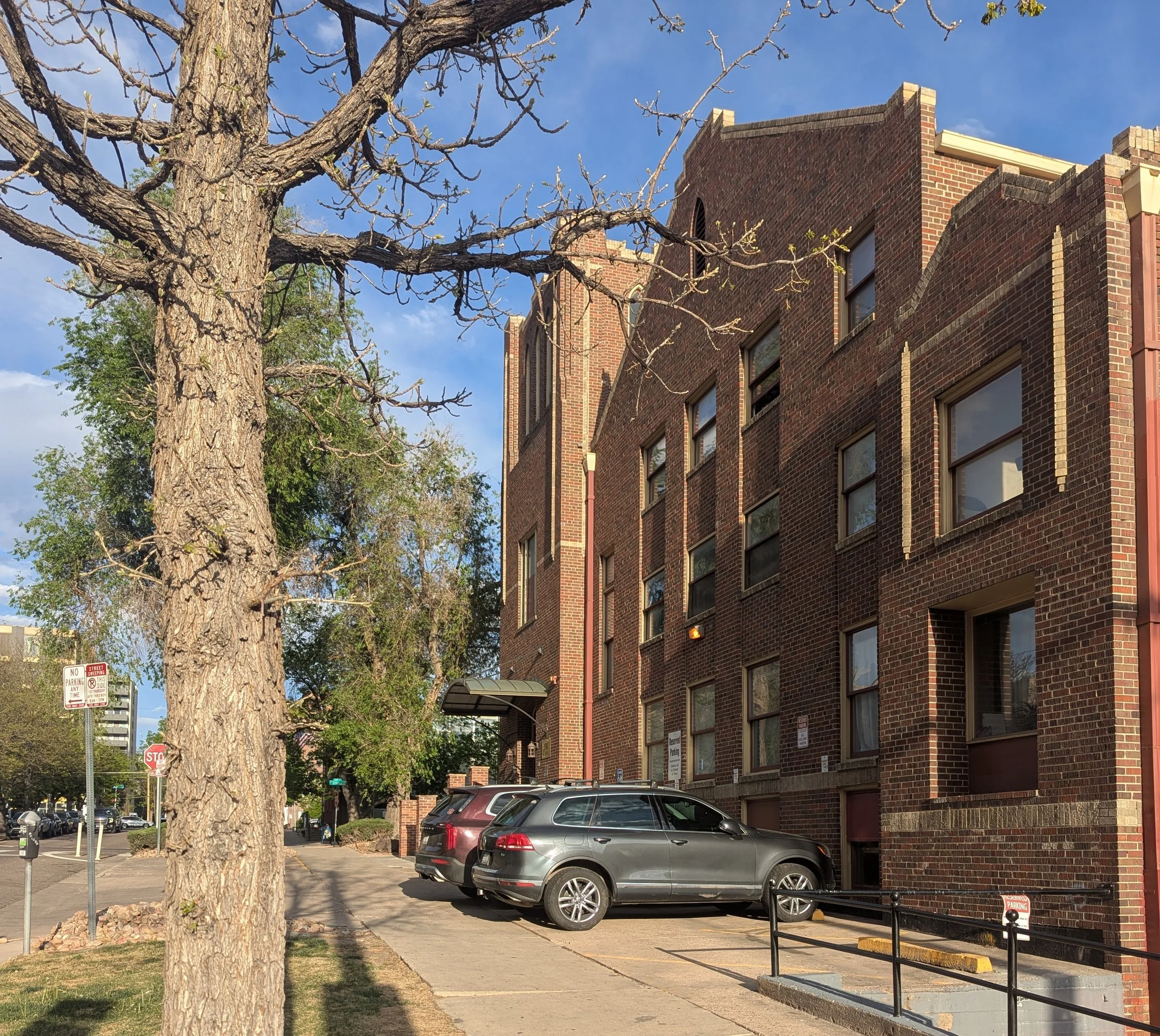 A city street with a brick building, a large leafless tree, parked cars, and parking signs, under a partly cloudy sky.