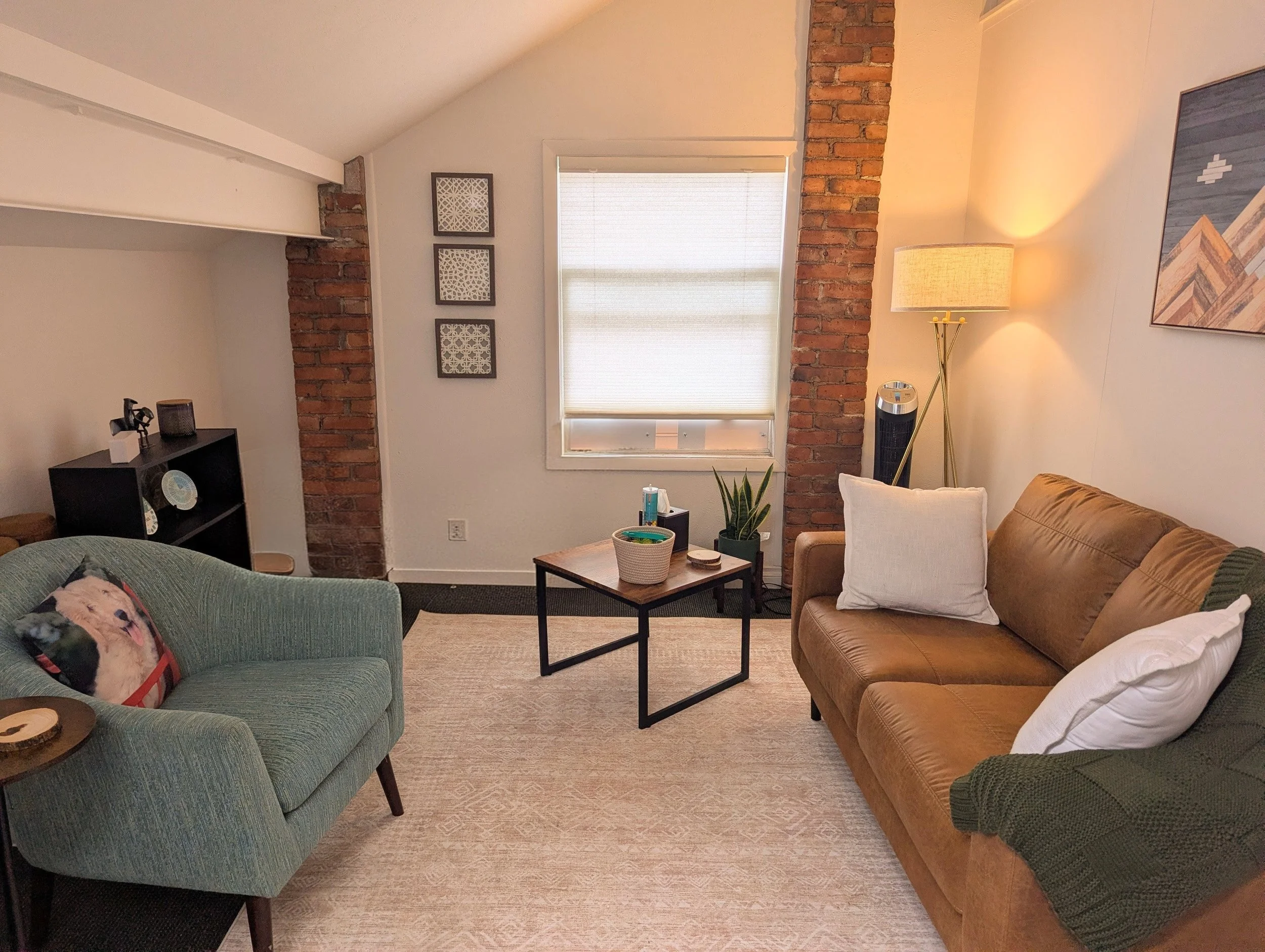 Living room with brick accents, a window with closed blinds, a floor lamp, a brown couch with white pillows, a green armchair with a dog pillow, and a small coffee table with plants and books.