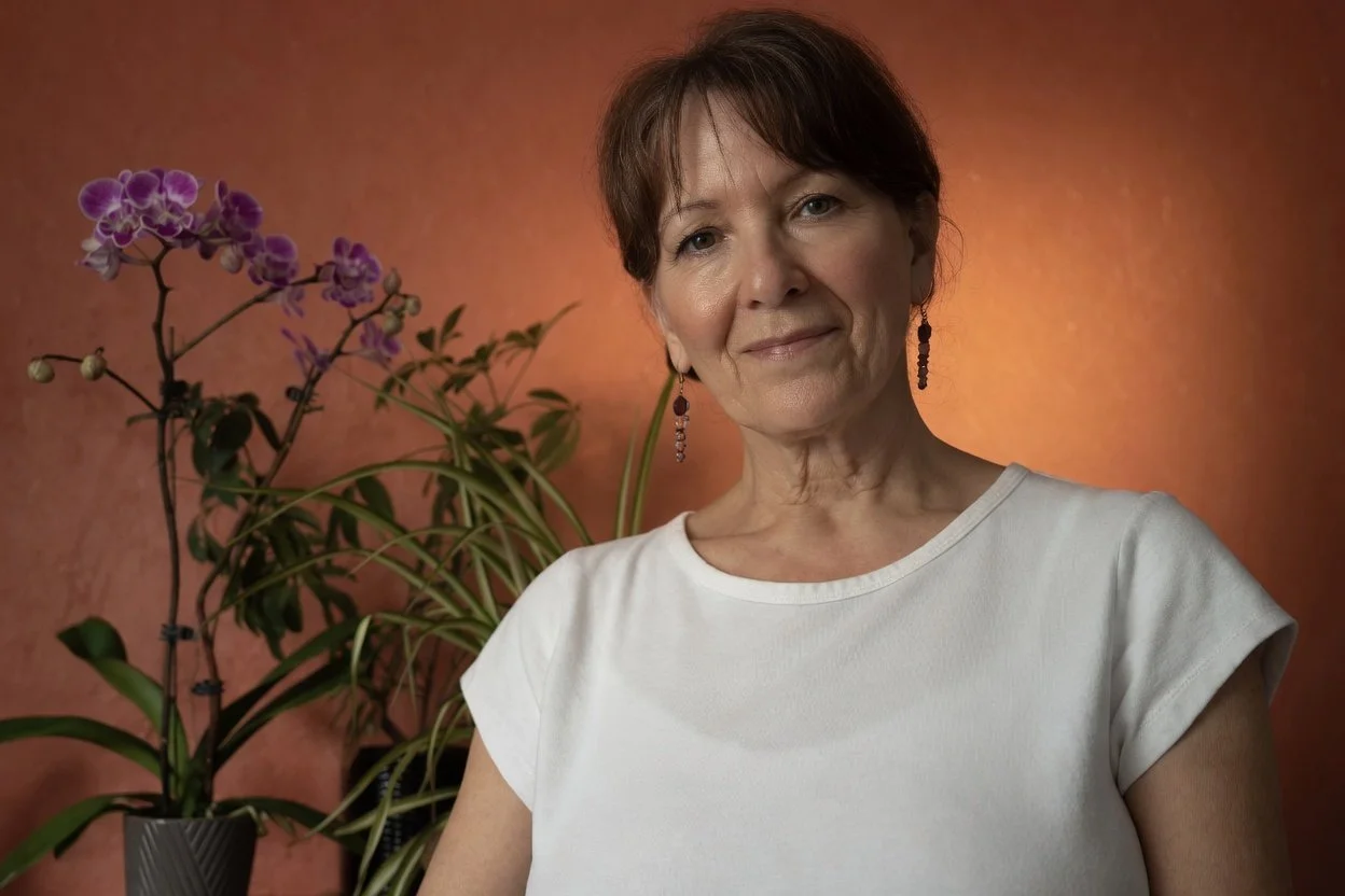 A middle-aged woman with short brown hair, wearing a white top and black earrings, stands in front of a tan backdrop with a potted purple orchid plant behind her.