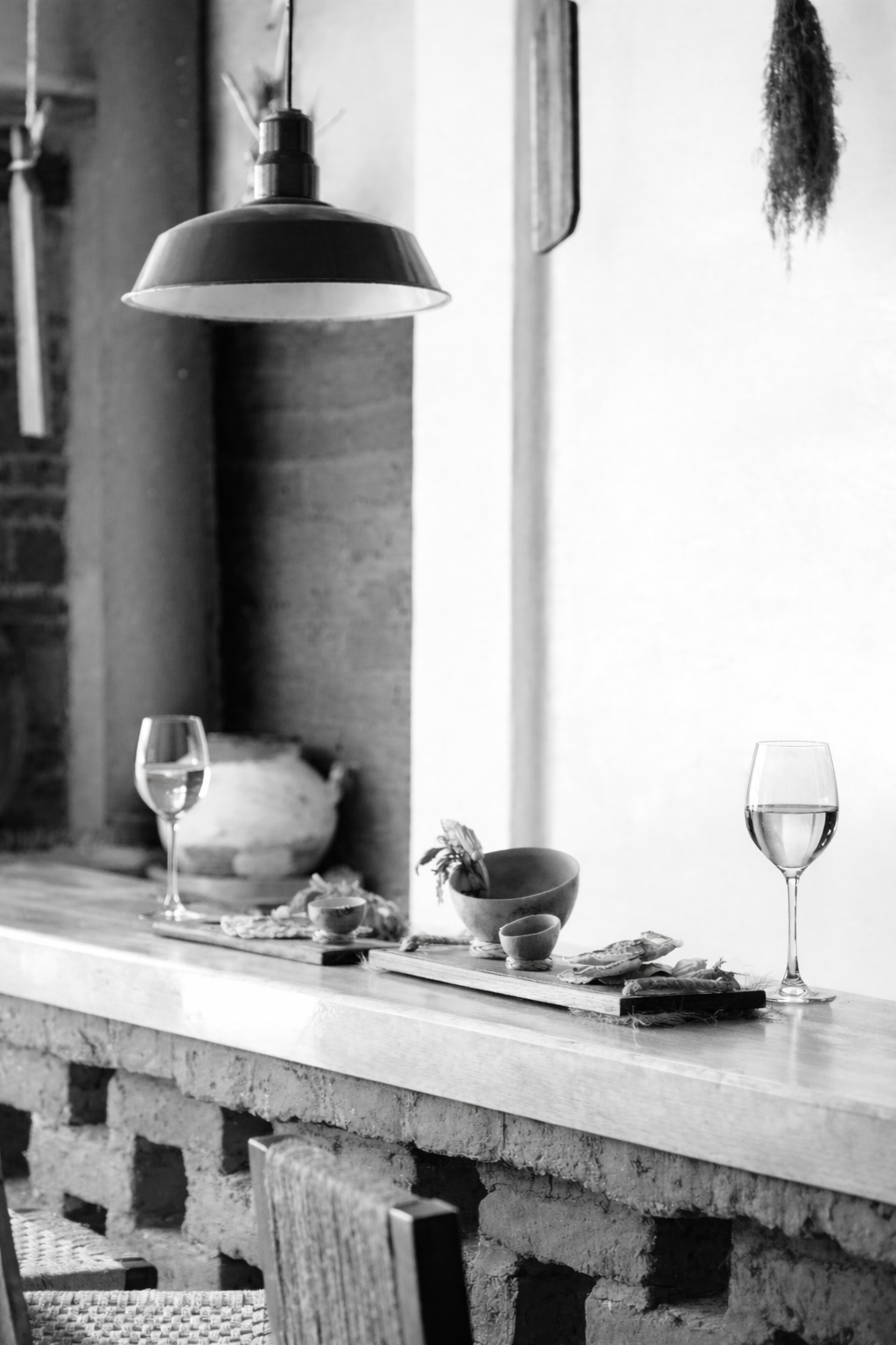Vista en blanco y negro de una mesa con vasos de vino, platos y decoraciones junto a una pared de ladrillos y una lámpara colgante arriba.