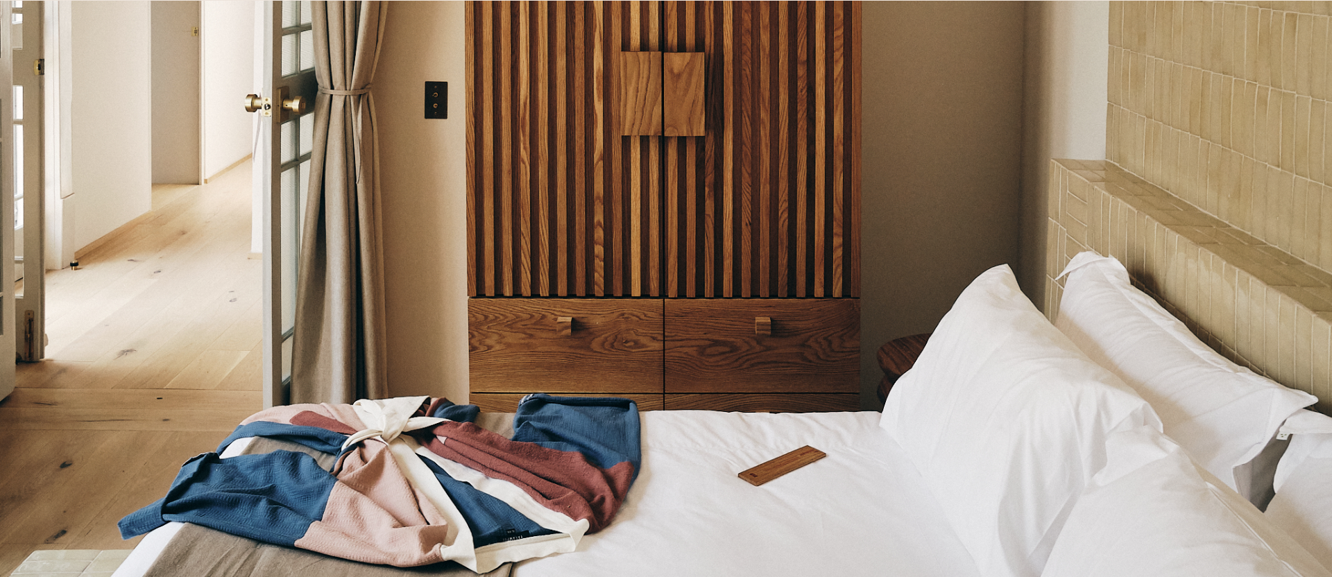 Cama con sábanas blancas y ropa doblada en colores rosa, azul y beige en un hotel, pared de ladrillos y armario de madera en el fondo, puerta abierta hacia un pasillo.
