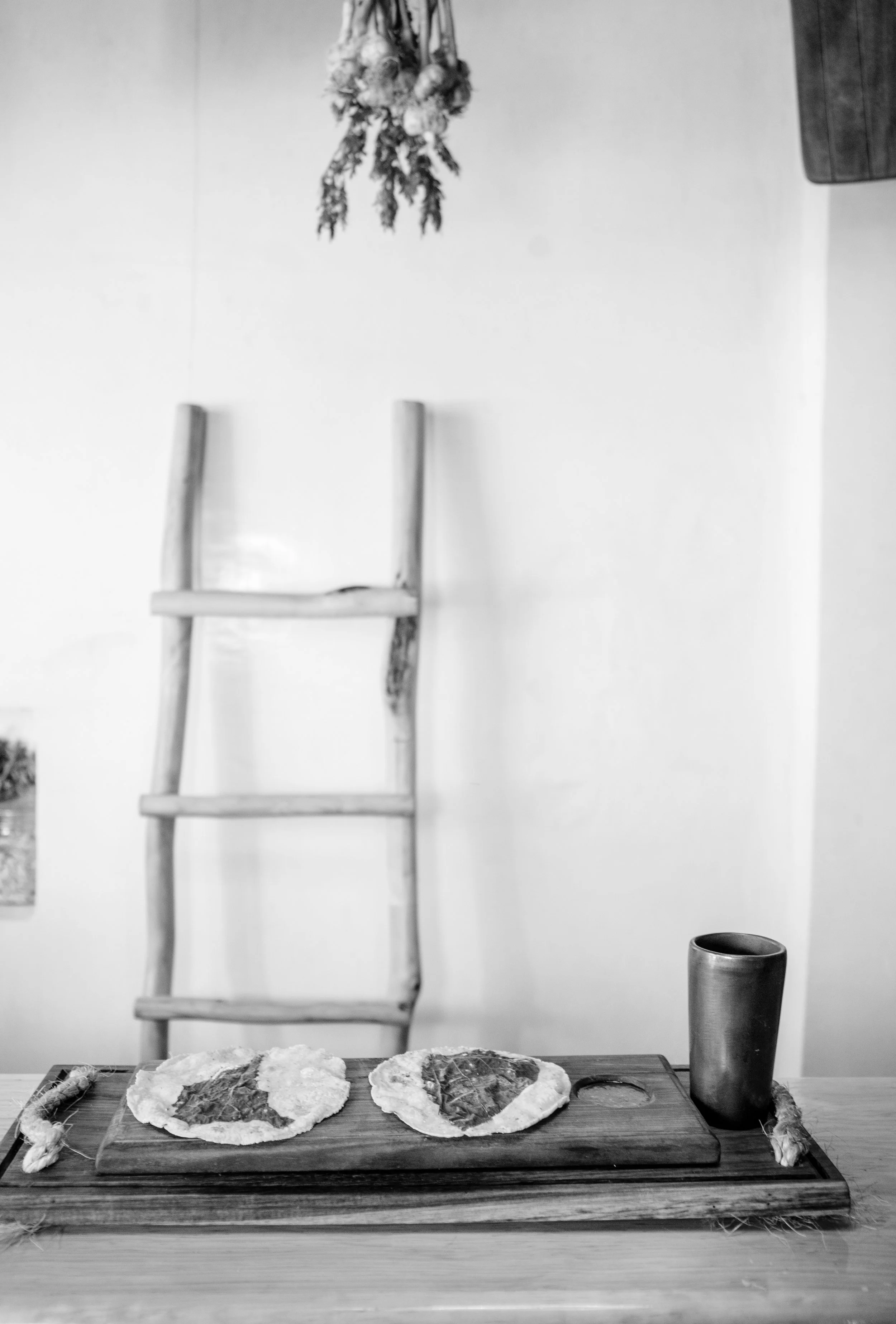 Mesa con dos tortillas y un vaso, pared blanca, escalera de madera colgando y plantas secas.