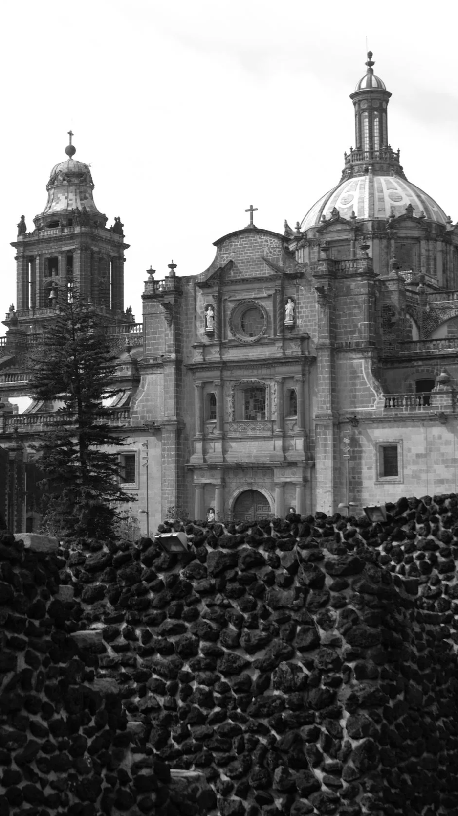 Una iglesia histórica de arquitectura colonial con cúpulas y torres, y un muro de piedra en primer plano.